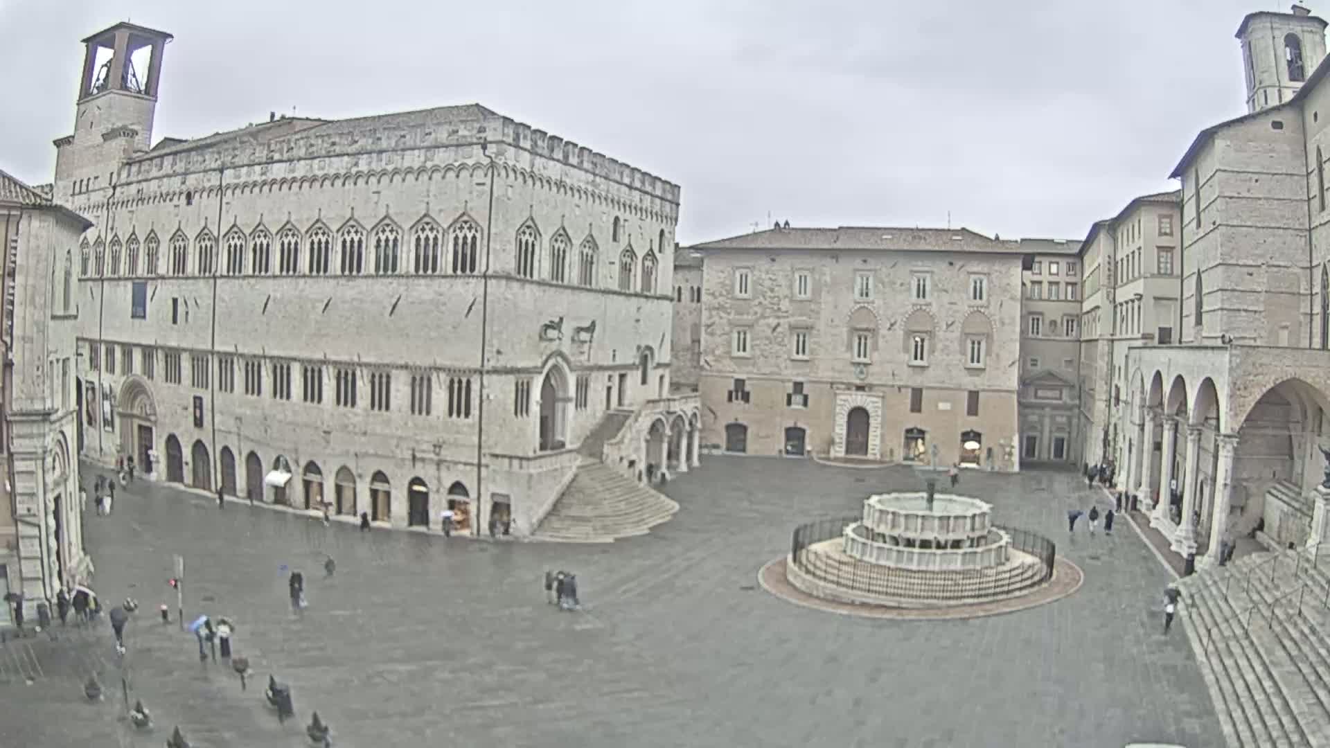 A large town square with several stone buildings, a fountain, and a few people walking around under an overcast sky.