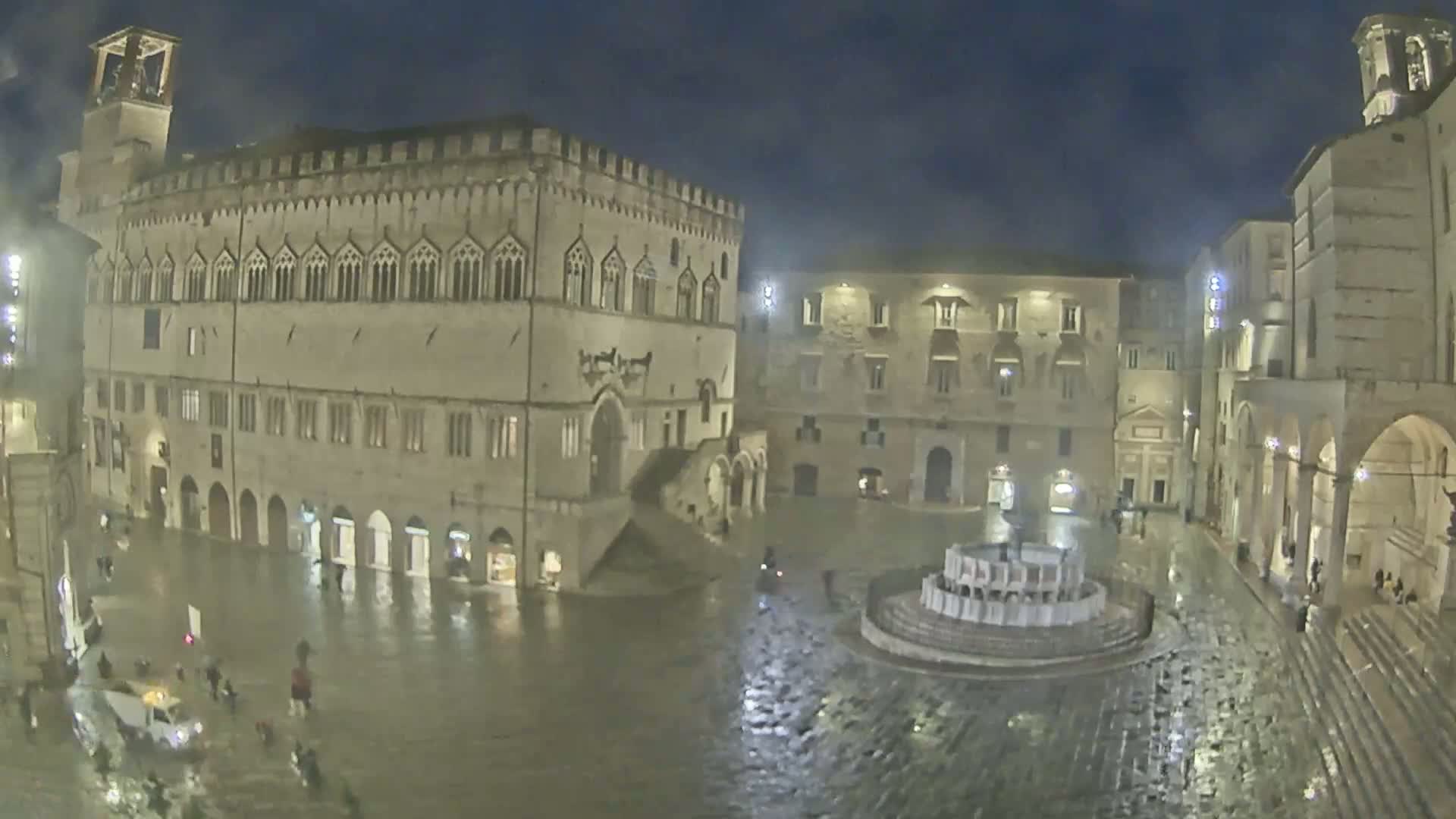 A large, mostly empty town square at night, surrounded by historic stone buildings, is flooded with rain.