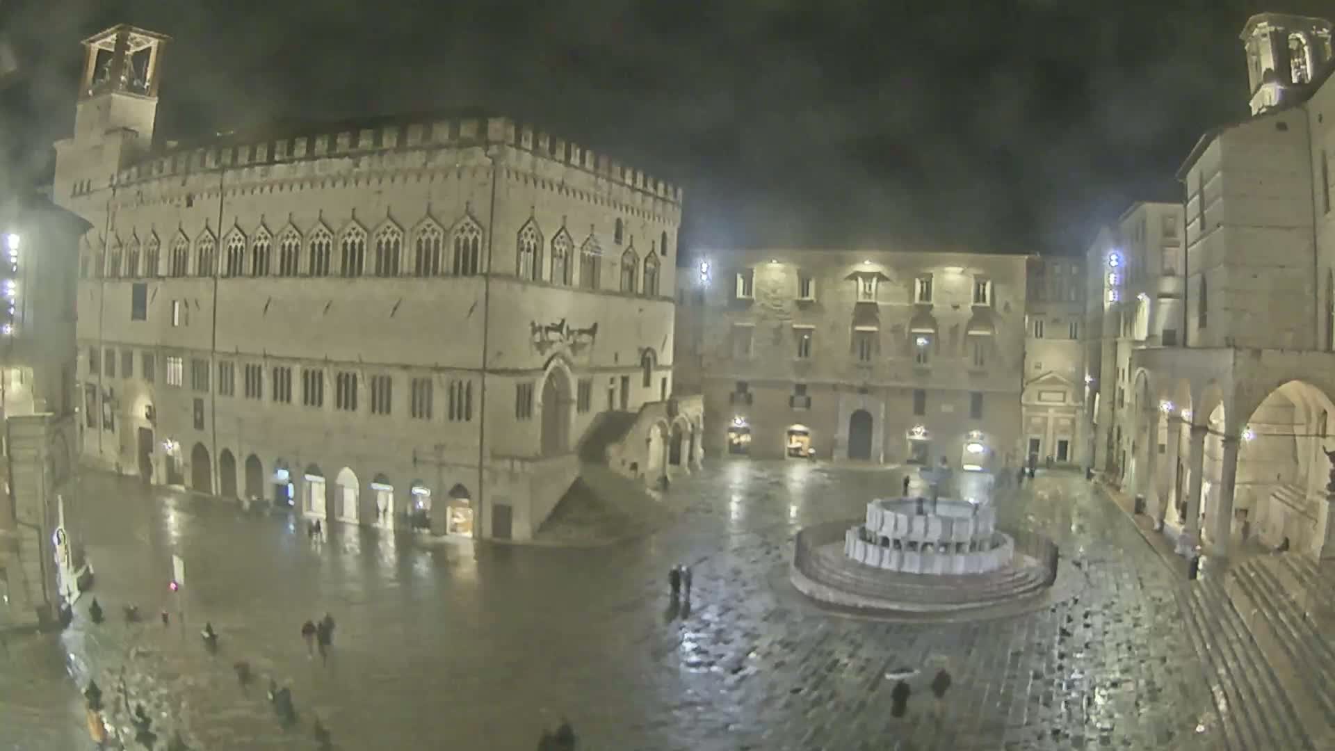 A nighttime, rain-slicked town square features a large, ornate fountain and several historic buildings.