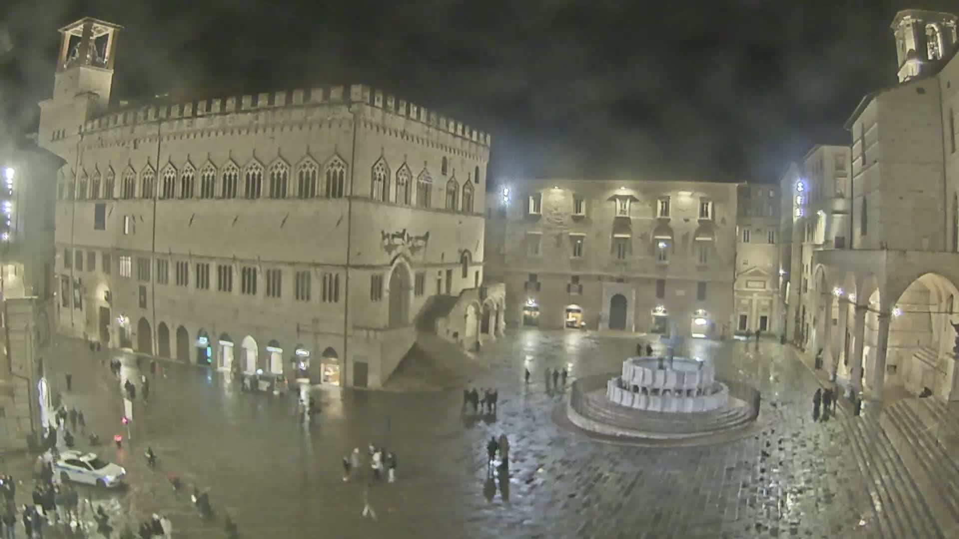 A rain-slicked town square at night, surrounded by large, illuminated stone buildings and a central fountain, features a few scattered pedestrians.