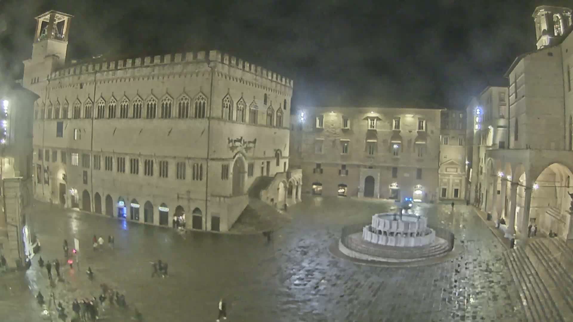 A rain-soaked town square at night, featuring a large stone building with arched windows and a central fountain.