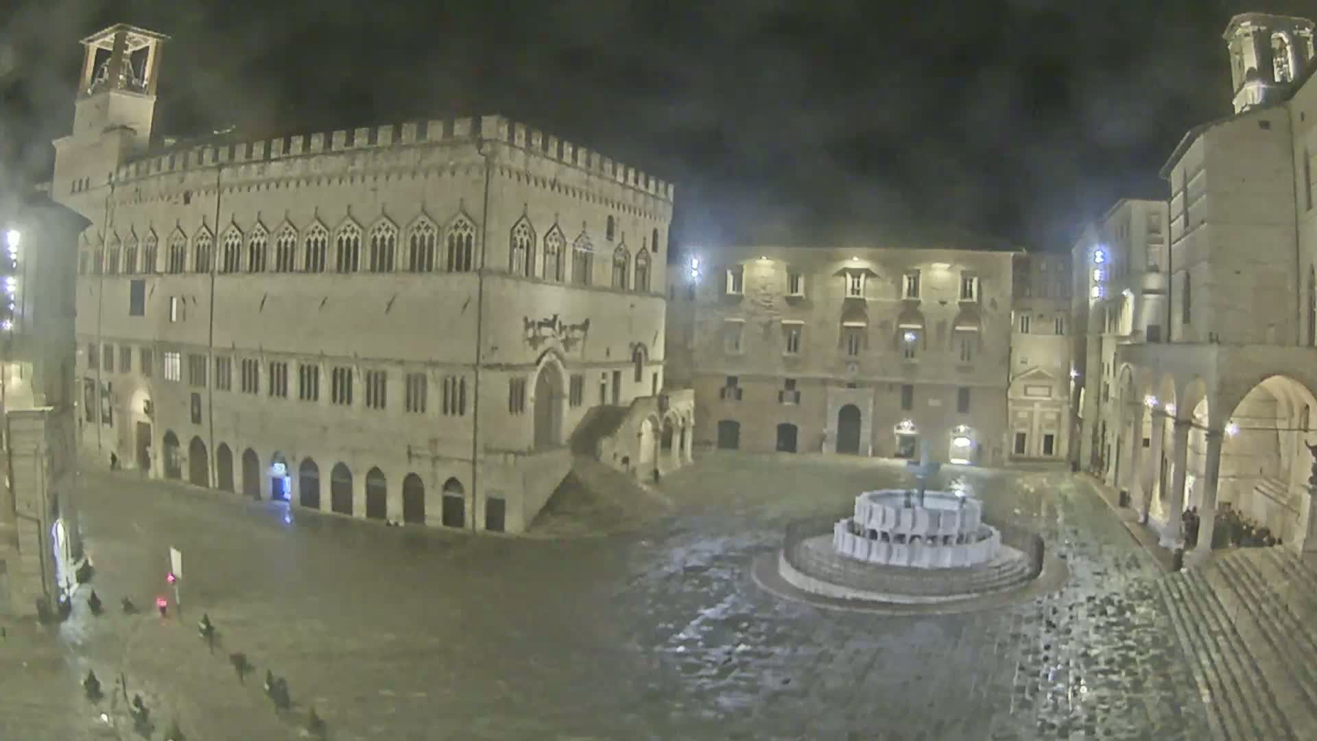 A large, mostly empty, stone plaza at night, lit by streetlights, features a central fountain and is surrounded by historic buildings under a dark, possibly rainy sky.