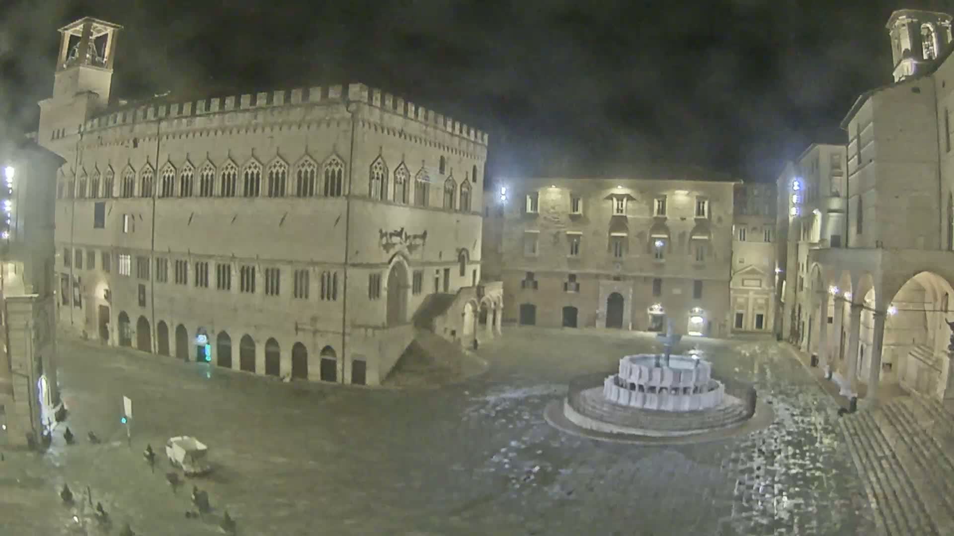 A nighttime view of a town square with a large stone building and fountain, under wet conditions.