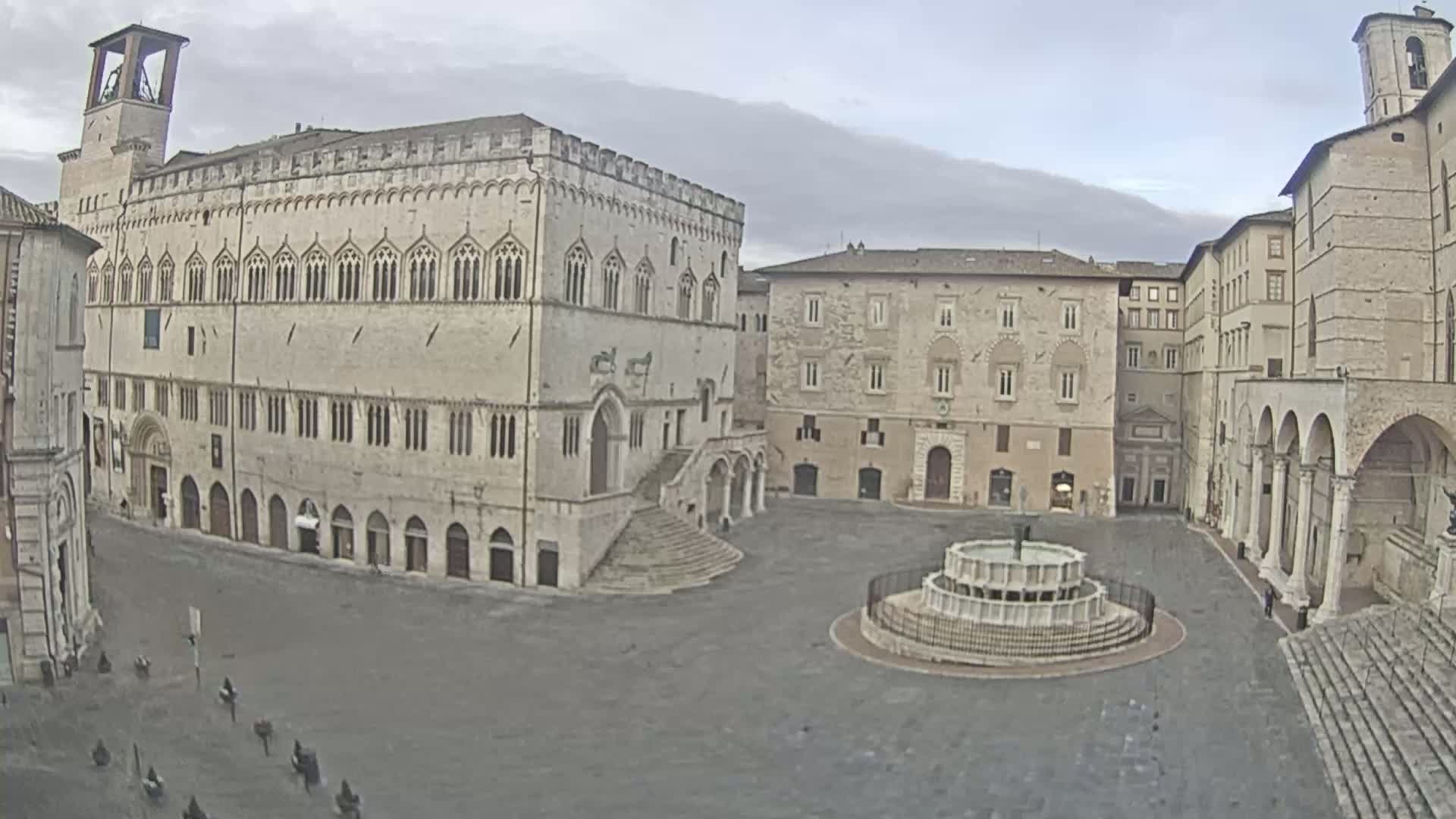 A large, empty town square with several stone buildings surrounds a circular fountain under an overcast sky.