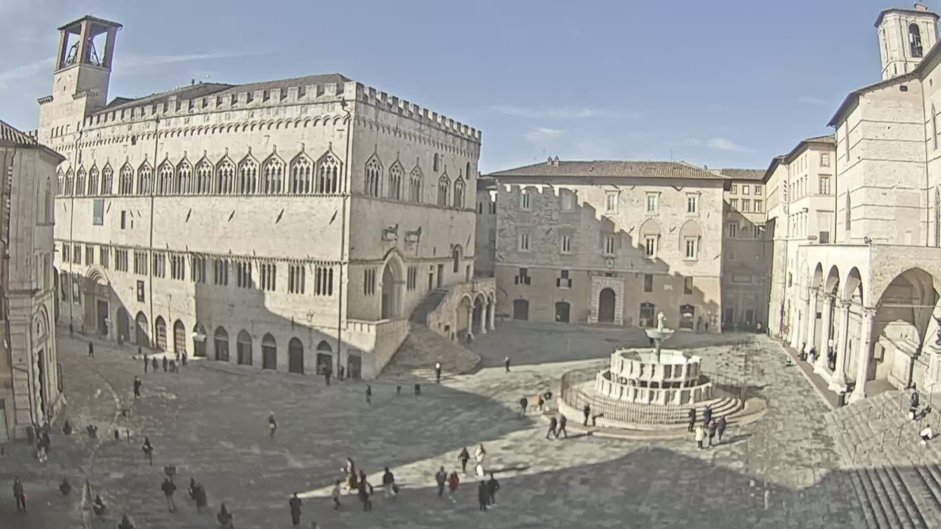 A large, paved town square with several historic stone buildings surrounding a central fountain, under a bright, sunny sky.
