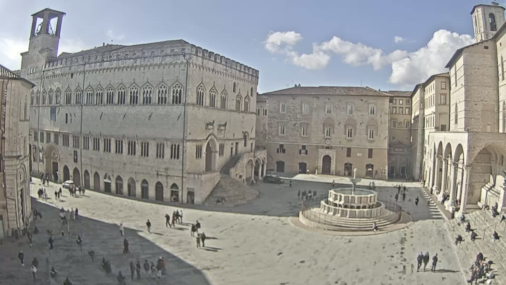 A large, sunlit town square with several stone buildings, a fountain in the center, and a scattering of people.