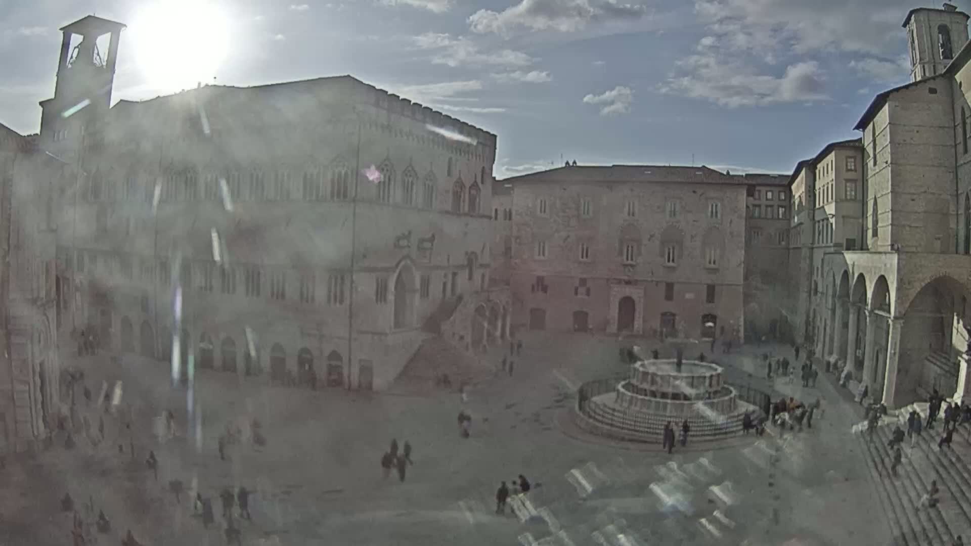 A sunny day reveals a large town square surrounded by historic stone buildings, a central fountain, and several people walking around.