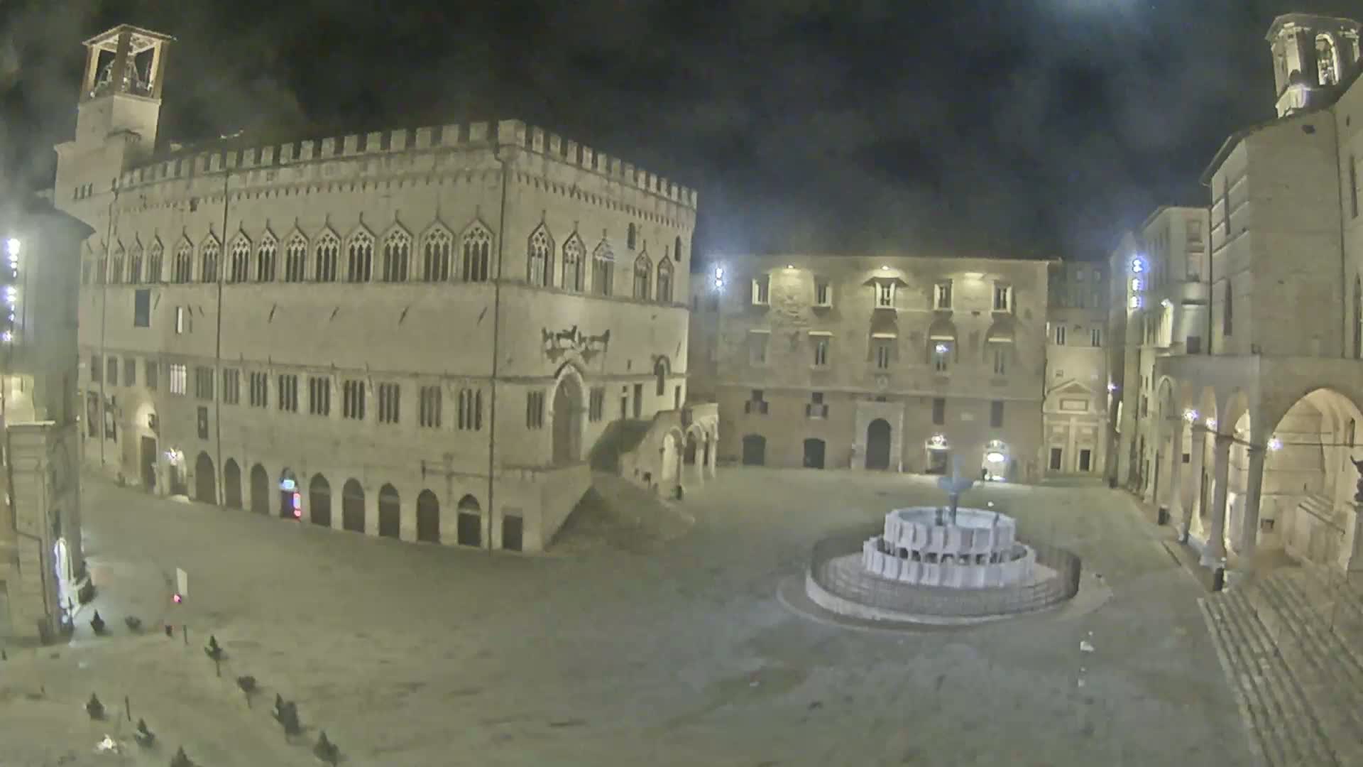 A mostly empty town square at night, featuring a large stone building and fountain, is lit by artificial light under a dark sky.