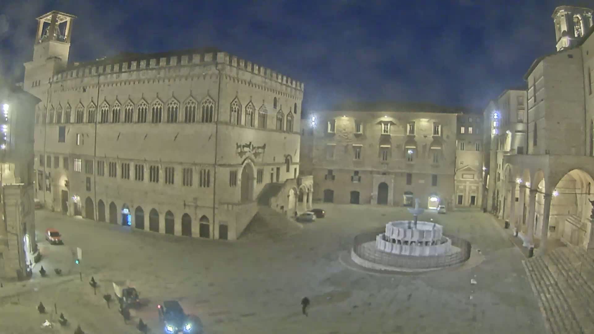 A nighttime view of a large, mostly empty town square with several historic buildings surrounding a fountain, under a dark sky.