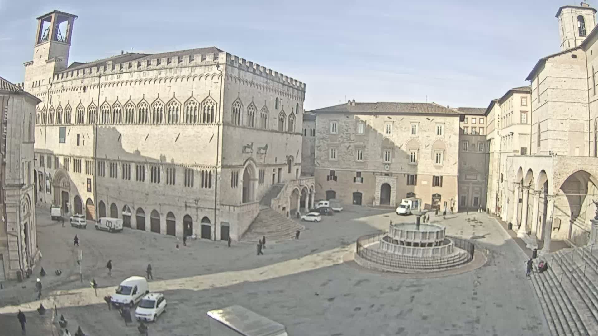 A town square, under a clear sunny sky, is surrounded by historic stone buildings, including a large palace with a staircase, and features a central fountain.