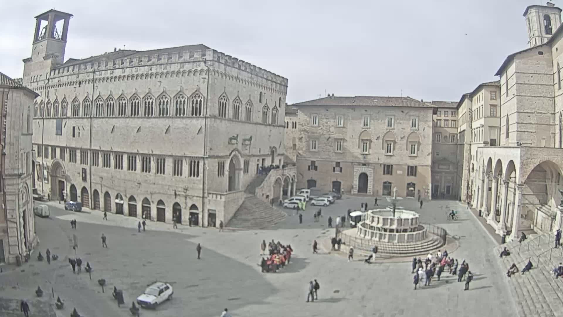A large, pale stone plaza is populated with a few people and cars, centered around a fountain, with several large, historic-looking buildings surrounding it under an overcast sky.