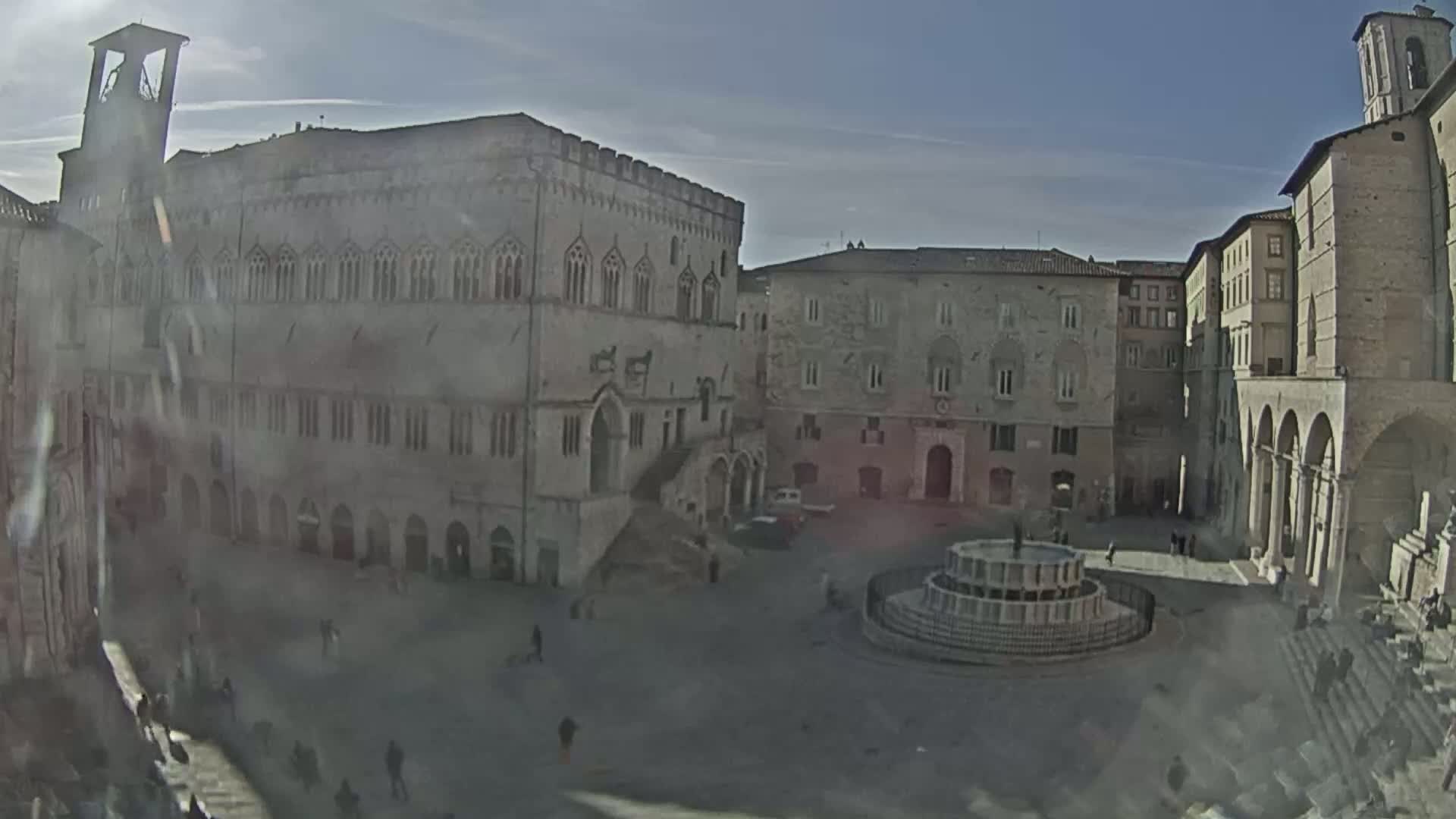 A sunny day reveals a large town square with several aged stone buildings surrounding a central fountain, a few people sparsely walking about.