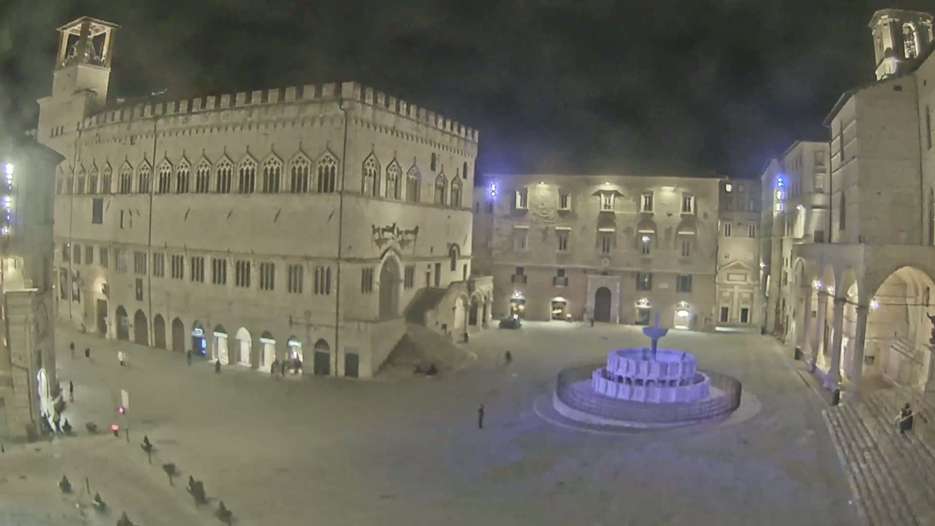 A large, mostly empty, stone plaza at night is illuminated, featuring a central fountain and surrounded by several historic buildings.