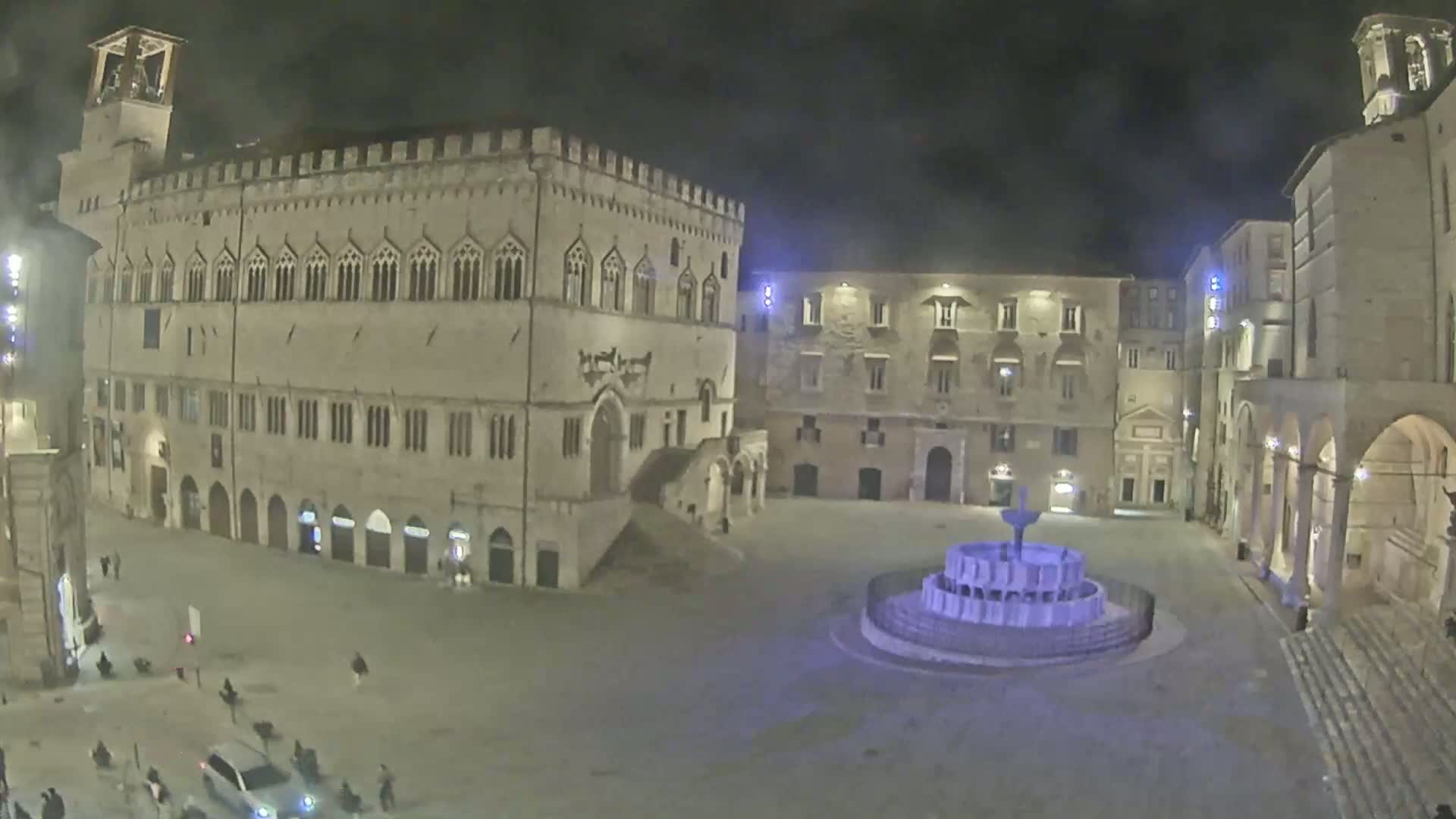 A nighttime, high-angle view of a town square with a large stone building, other buildings surrounding it, and a fountain in the center.