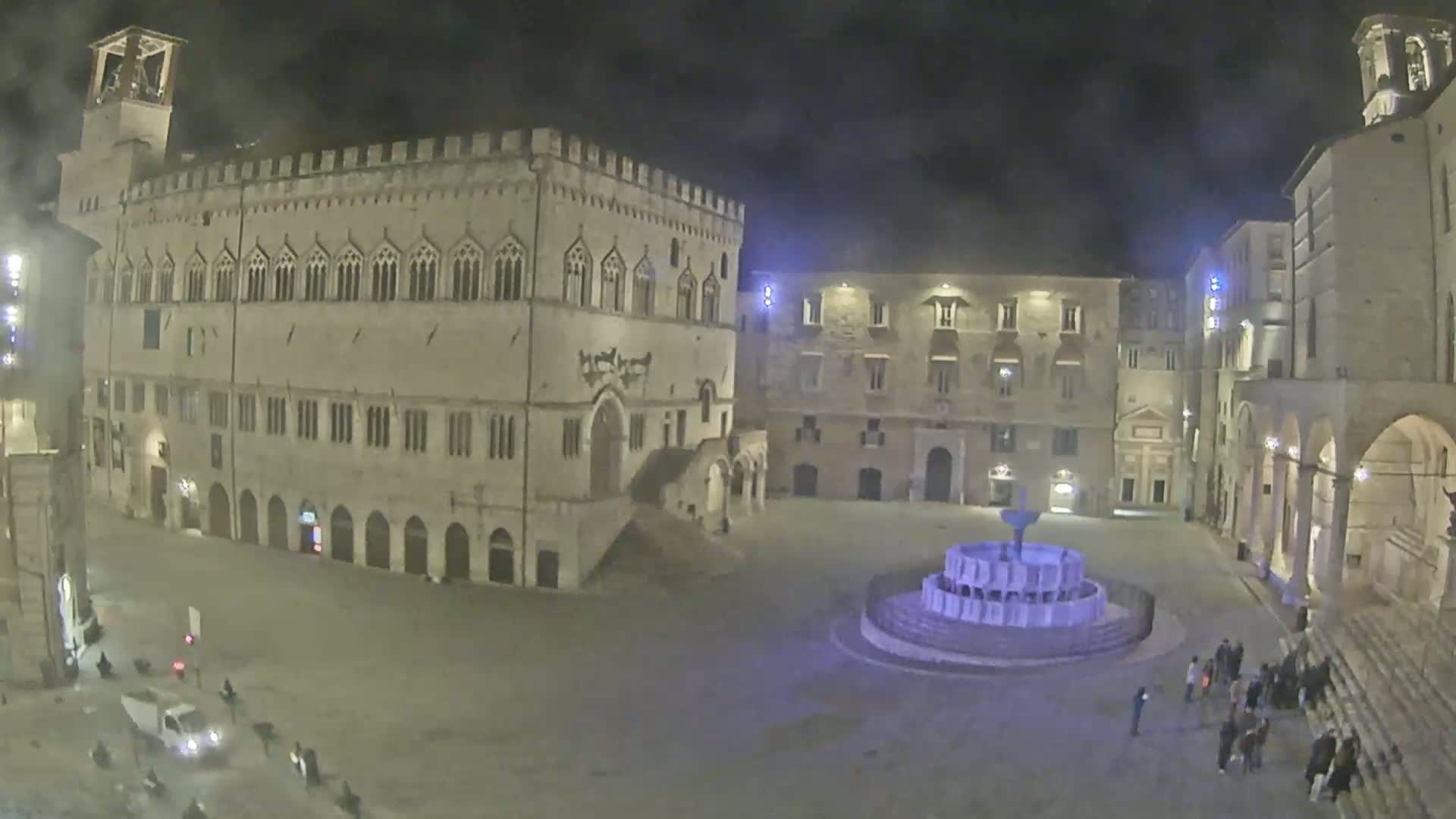 A nighttime view of a town square with several stone buildings surrounding a central fountain, under a dark sky.