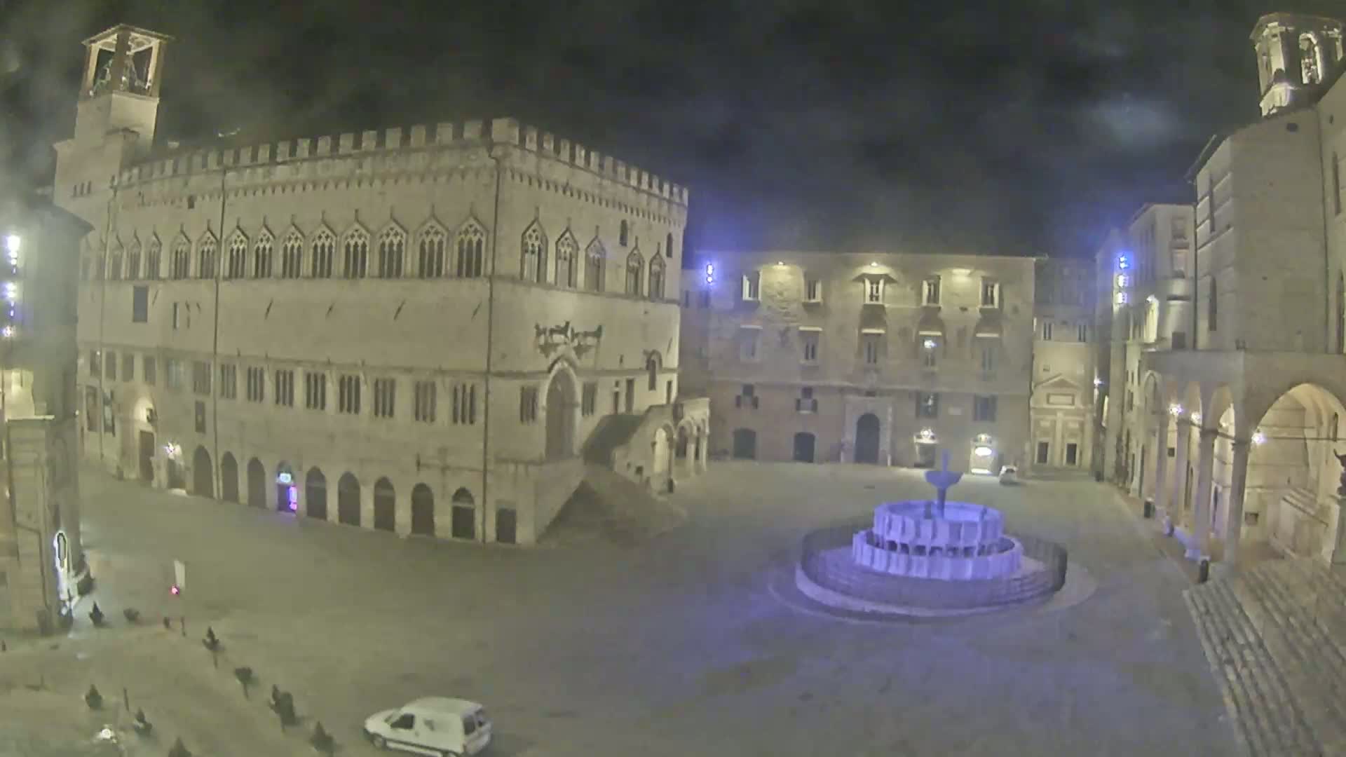 A nighttime view of a mostly empty town square, centered around a fountain, surrounded by ancient stone buildings under a cloudy sky.