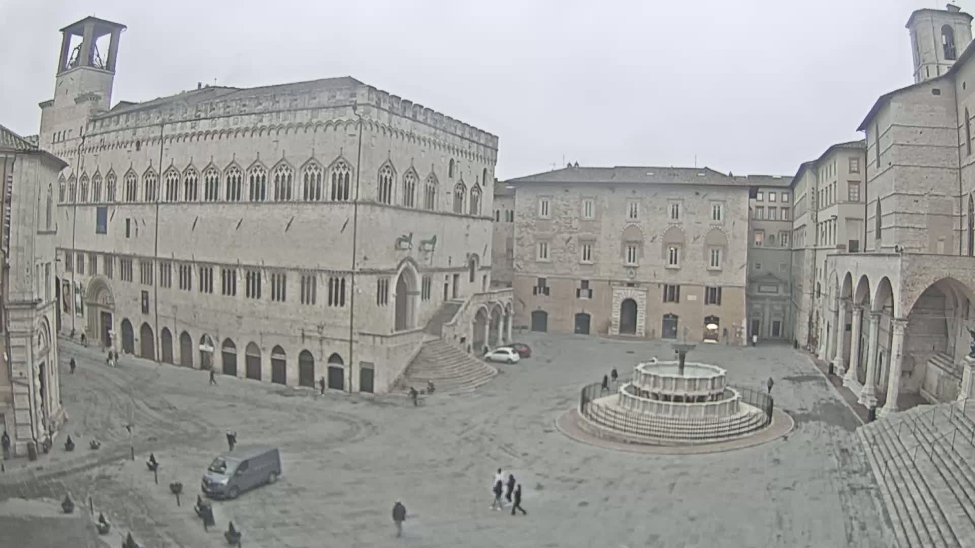 A large, stone plaza with a fountain in the center, surrounded by historic buildings under an overcast sky.
