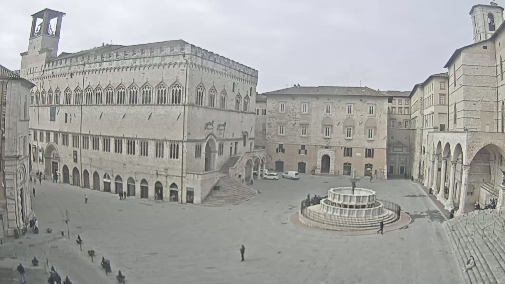A large, light-colored stone plaza with a fountain, surrounded by medieval-style buildings, under an overcast sky.