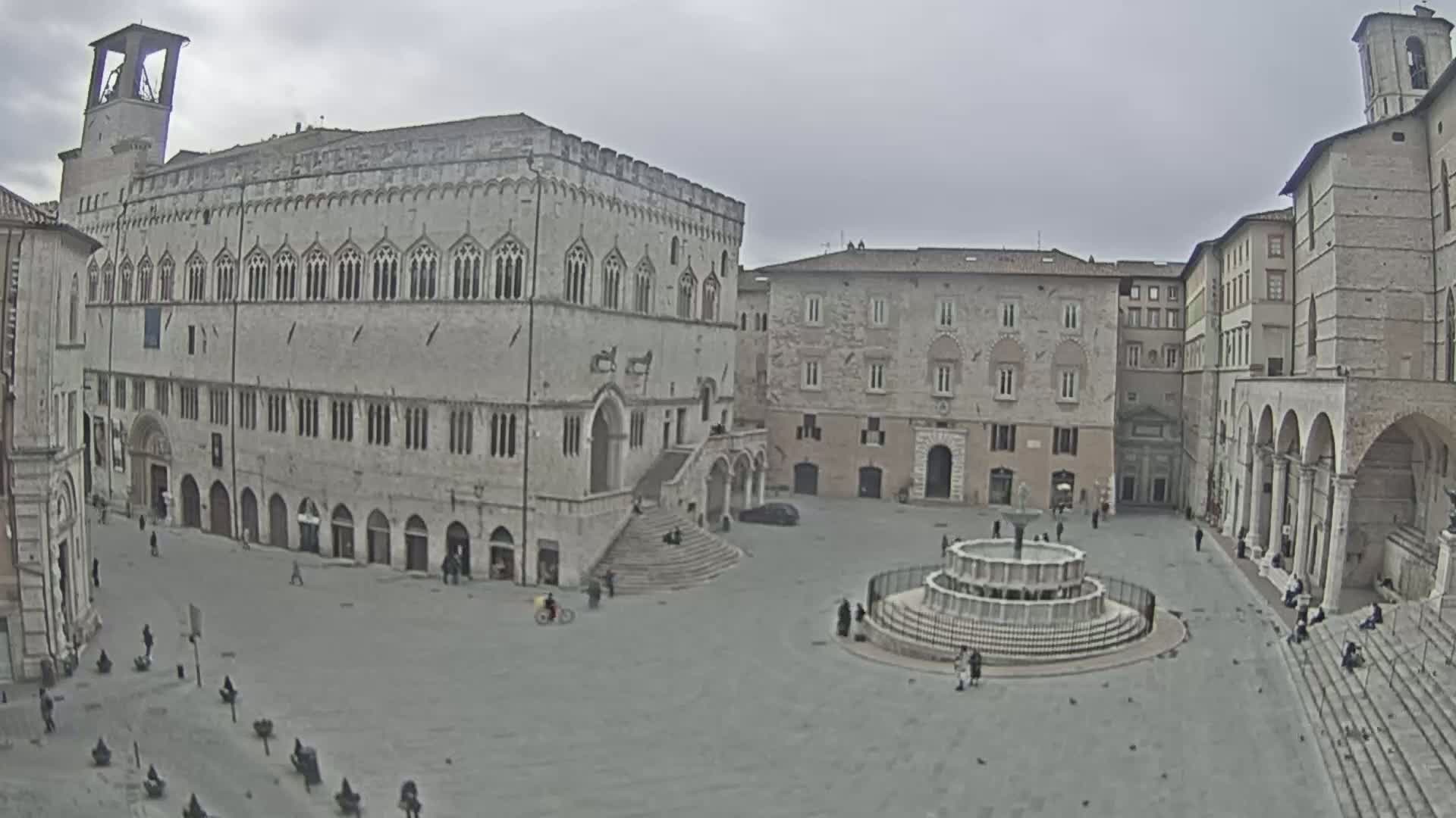 A large, mostly empty town square with a fountain at its center is surrounded by several medieval-style buildings under an overcast sky.