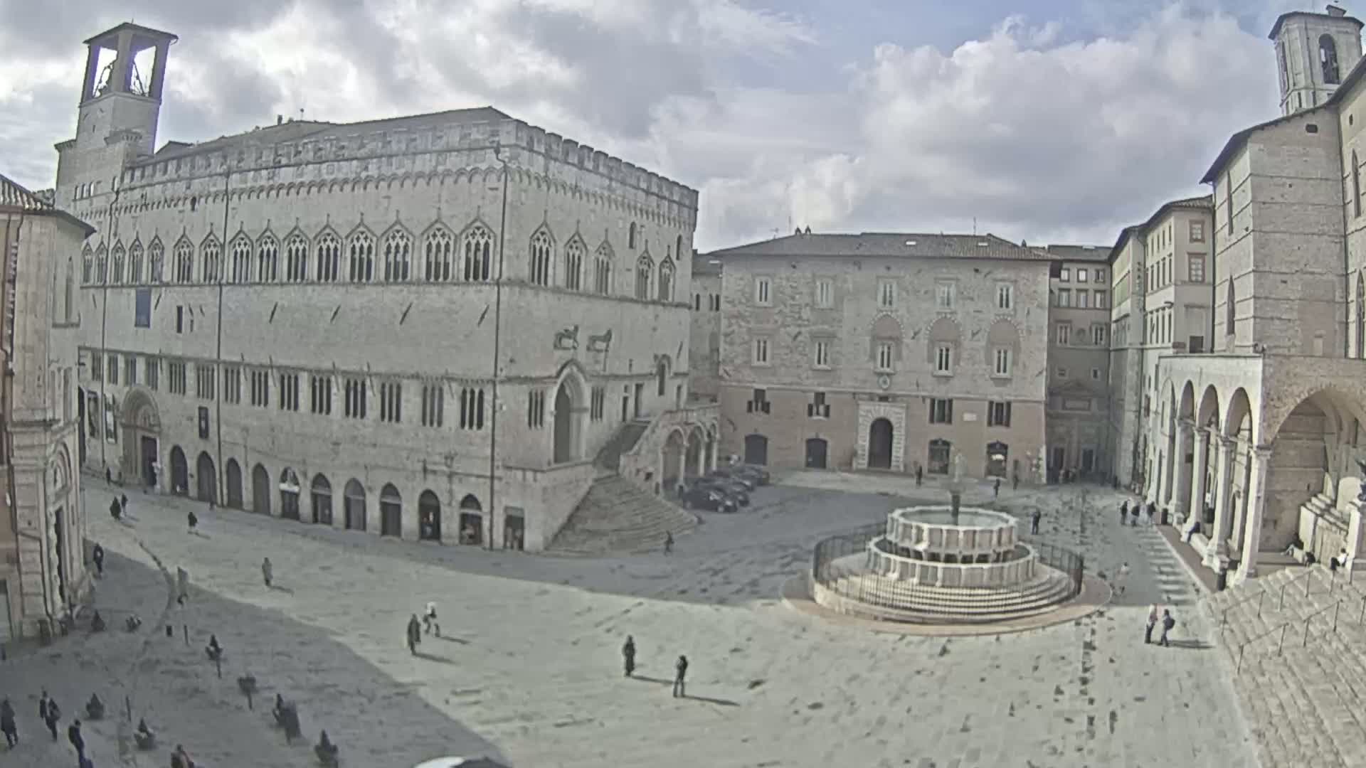 A large, stone plaza with a fountain in the center, surrounded by historic buildings under a cloudy sky.