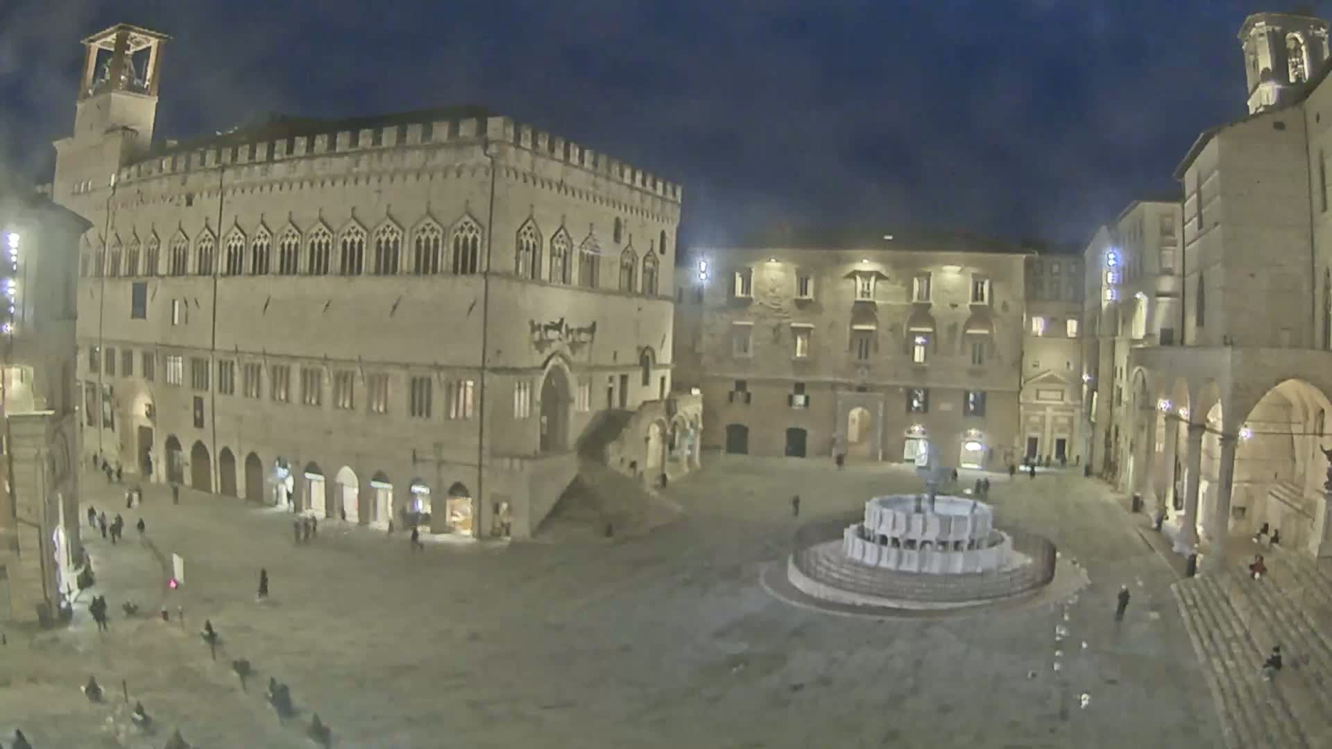 A nighttime view of a large, sparsely populated town square with a fountain at its center, surrounded by historic stone buildings under a dark sky.