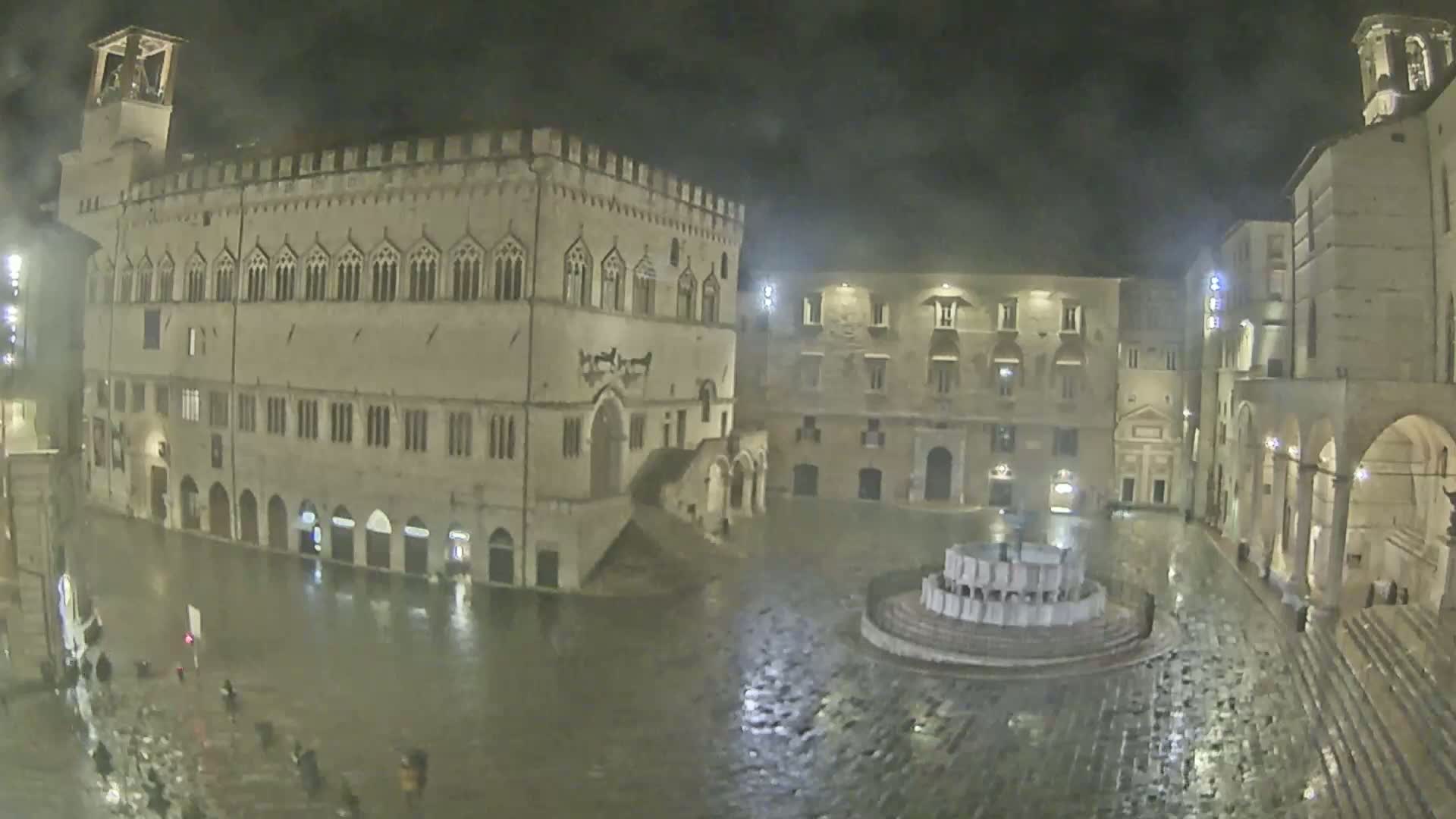 A large, flood-lit town square, partially flooded with rainwater, is surrounded by medieval-style buildings at night.