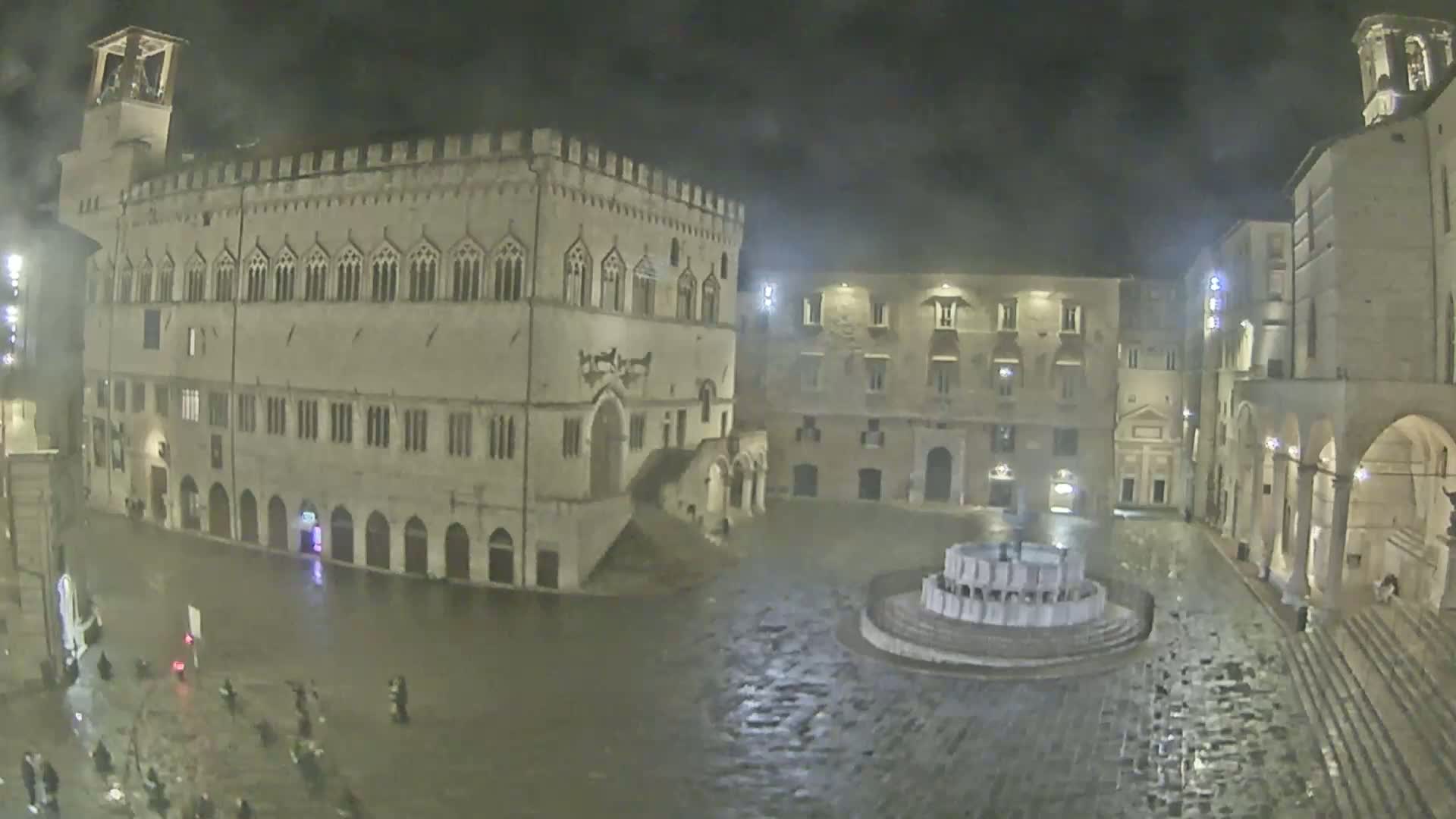 A nighttime, rain-soaked town square features a large stone building and several smaller buildings surrounding a fountain, with a few people walking through the square.