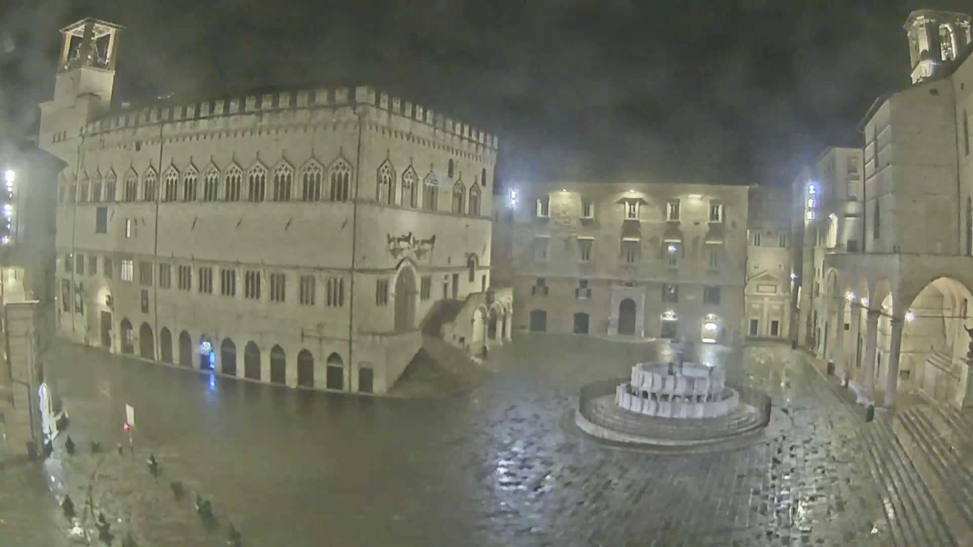 A floodlit, stone plaza at night, partially flooded with rain, is centered around a fountain and surrounded by historic buildings.