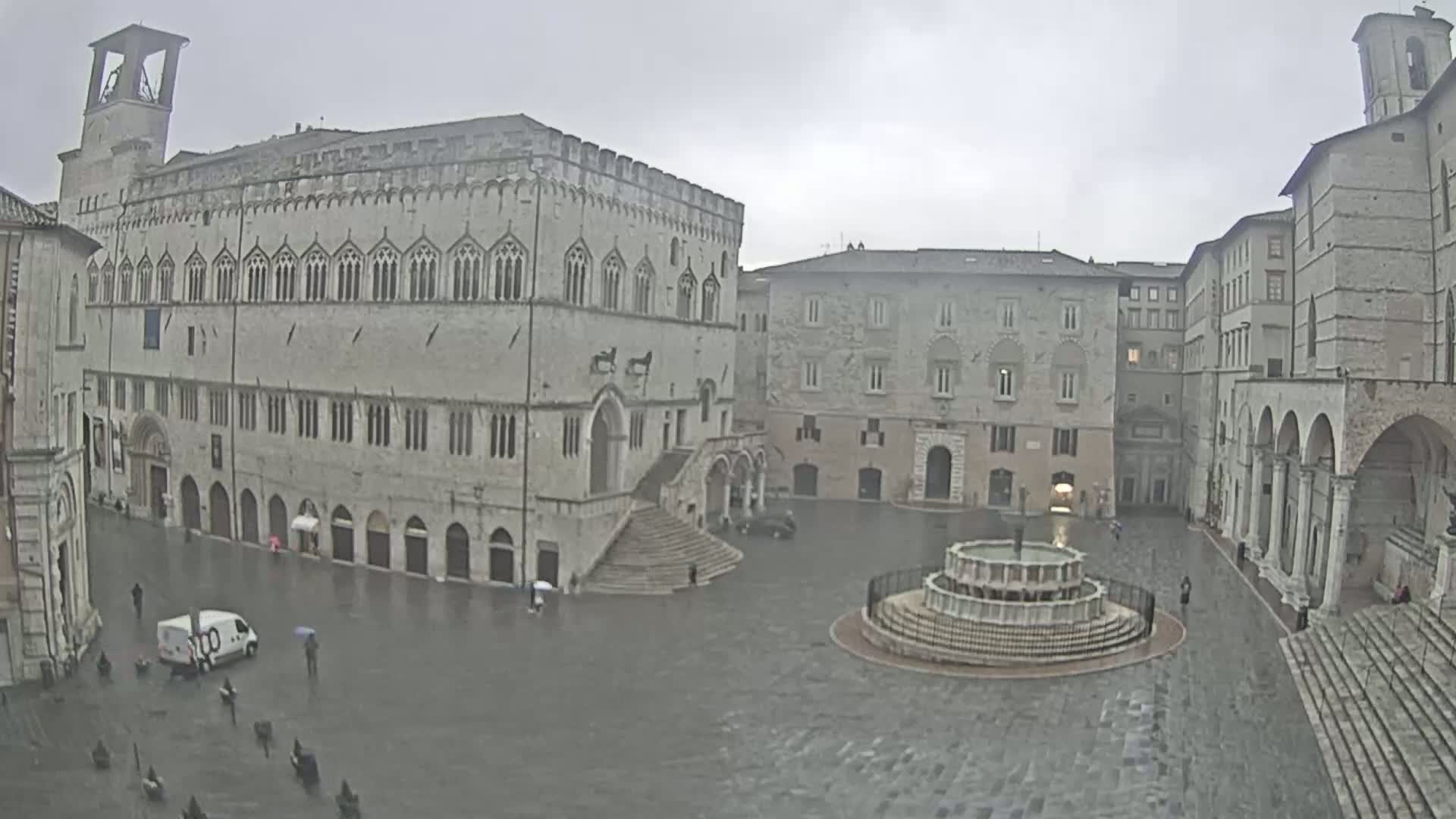 A large, stone piazza with a fountain in the center is surrounded by historic buildings under an overcast, rainy sky.