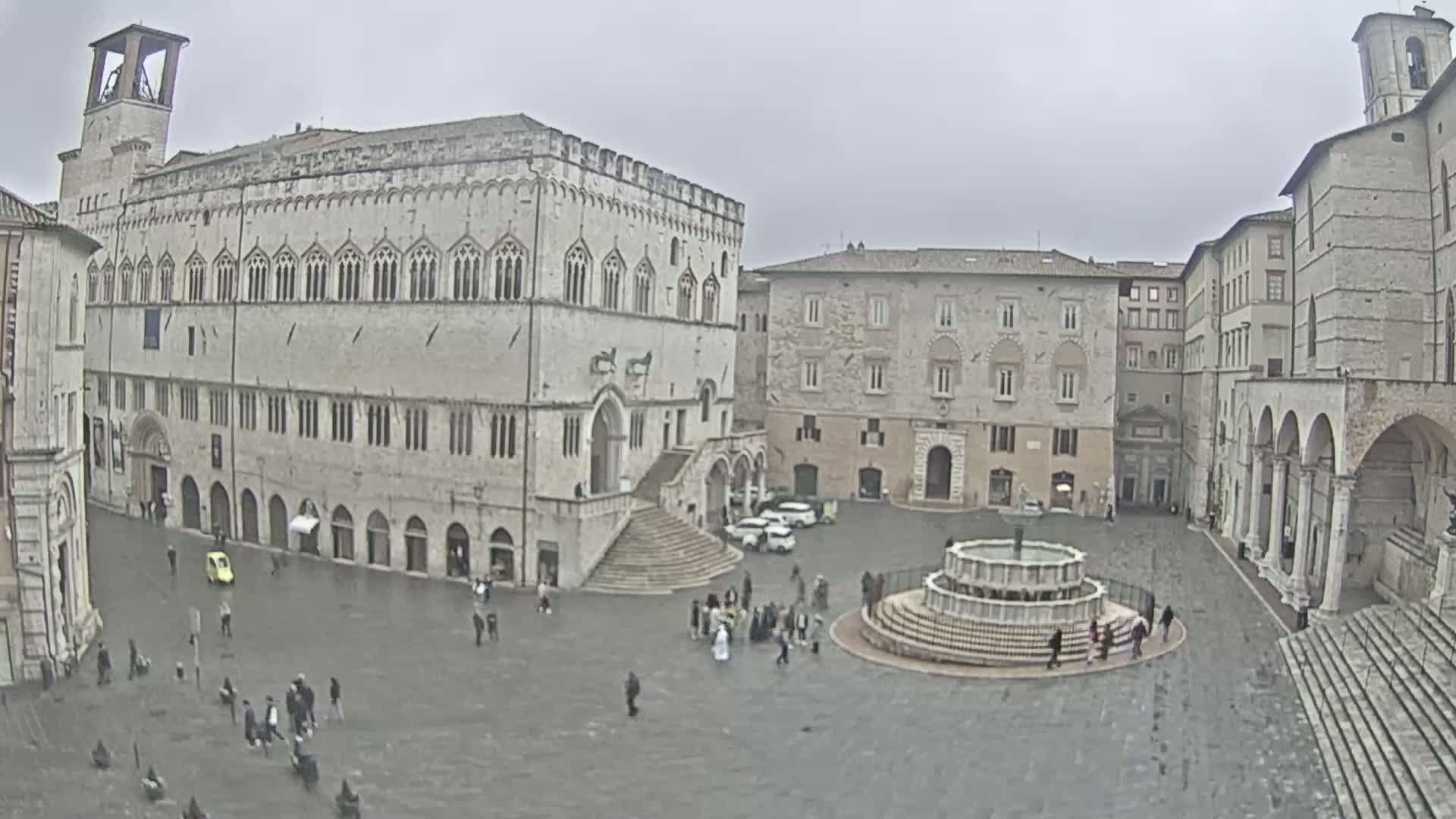A large, stone town square with a fountain at its center is surrounded by several historical buildings under an overcast sky.