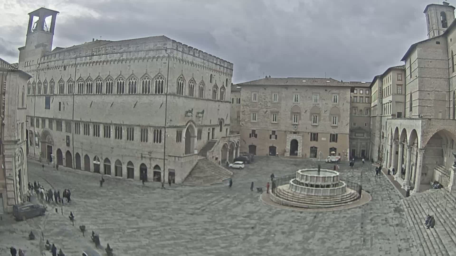 A large town square with several stone buildings, a fountain, and a few people, under an overcast sky.