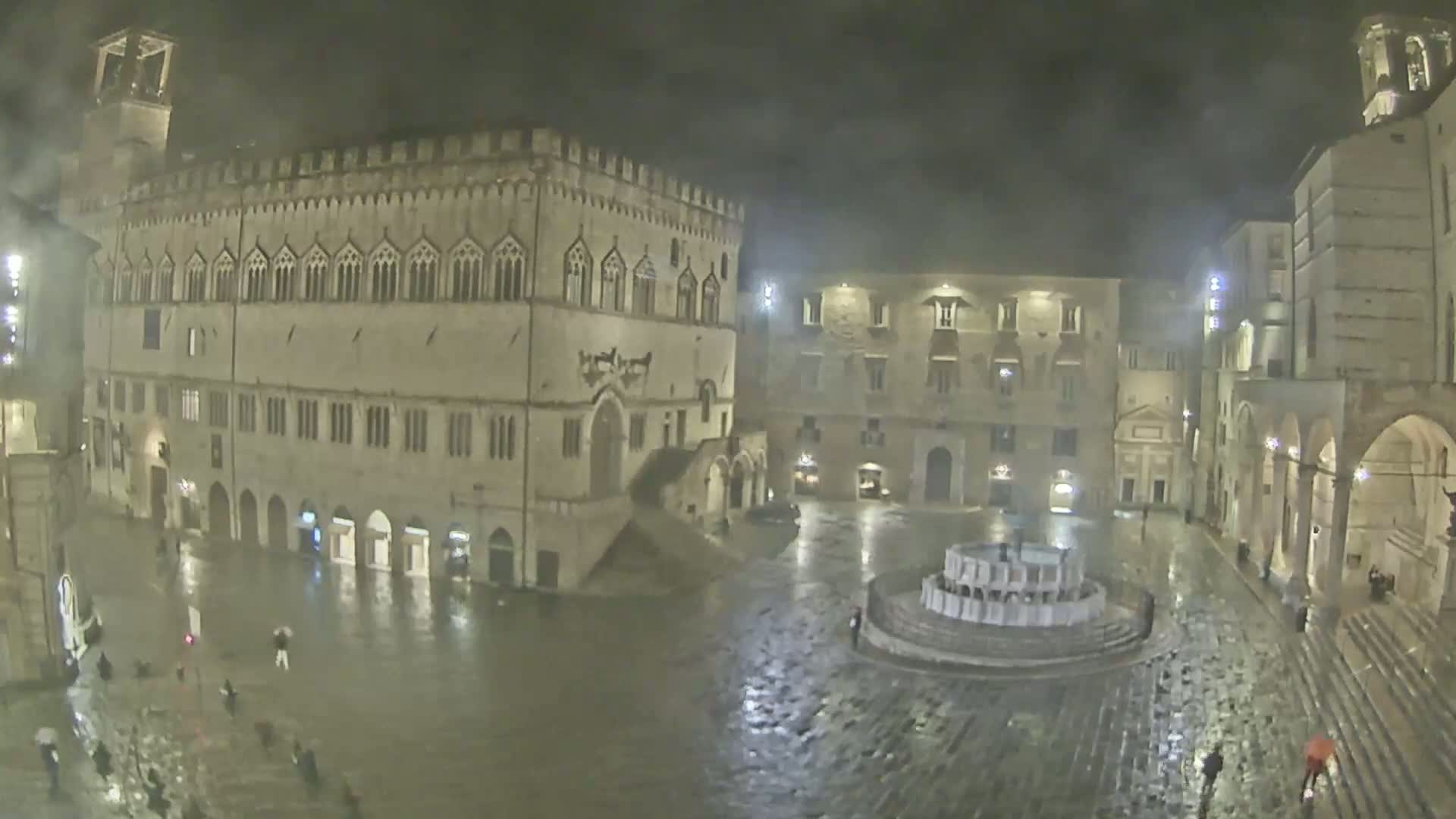 A rain-soaked town square at night, featuring a large stone building and a fountain, is sparsely populated with pedestrians.