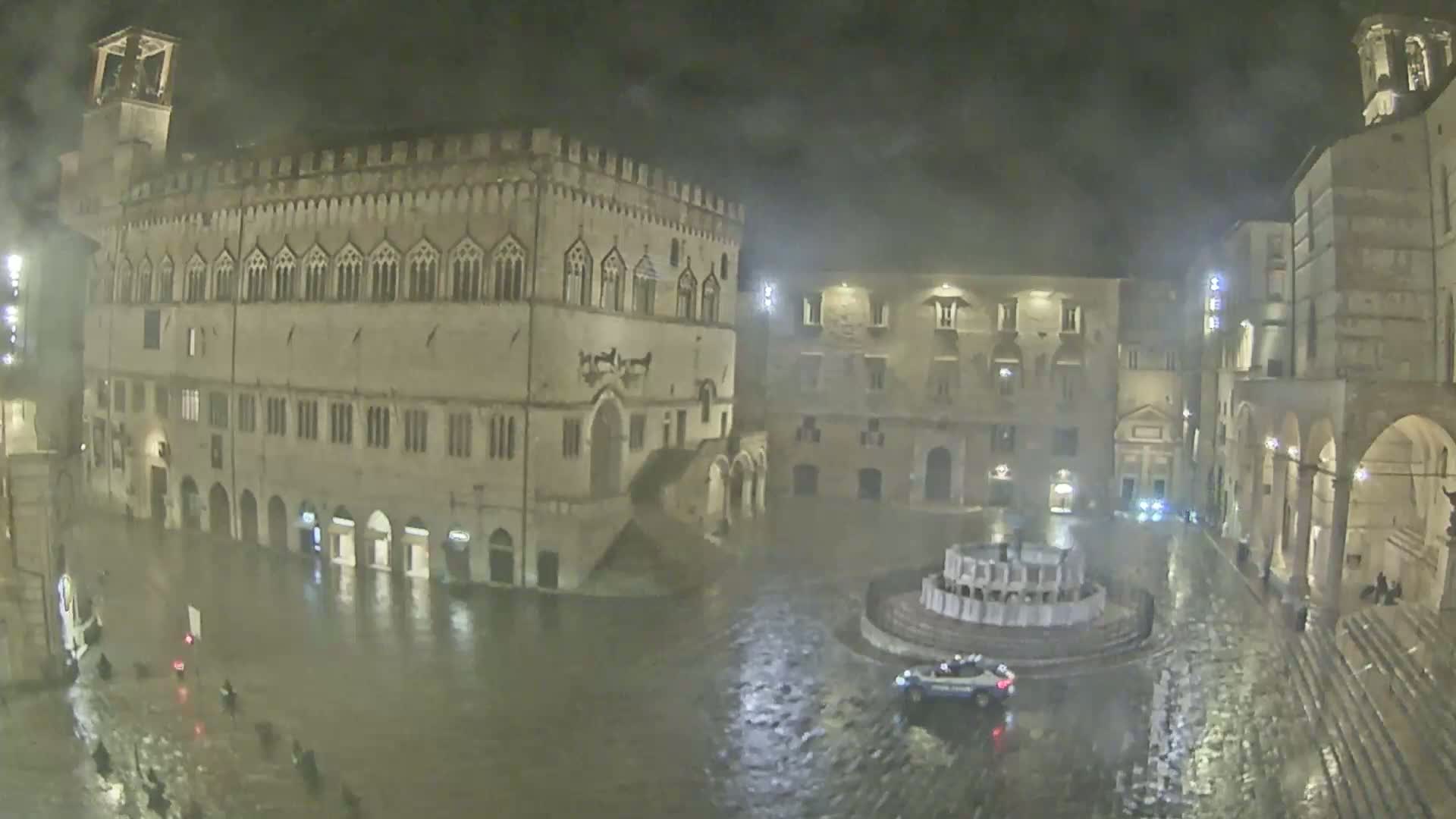 A floodlit town square, mostly submerged in rainwater at night, features several historical buildings surrounding a circular fountain, with a single vehicle visible in the square.