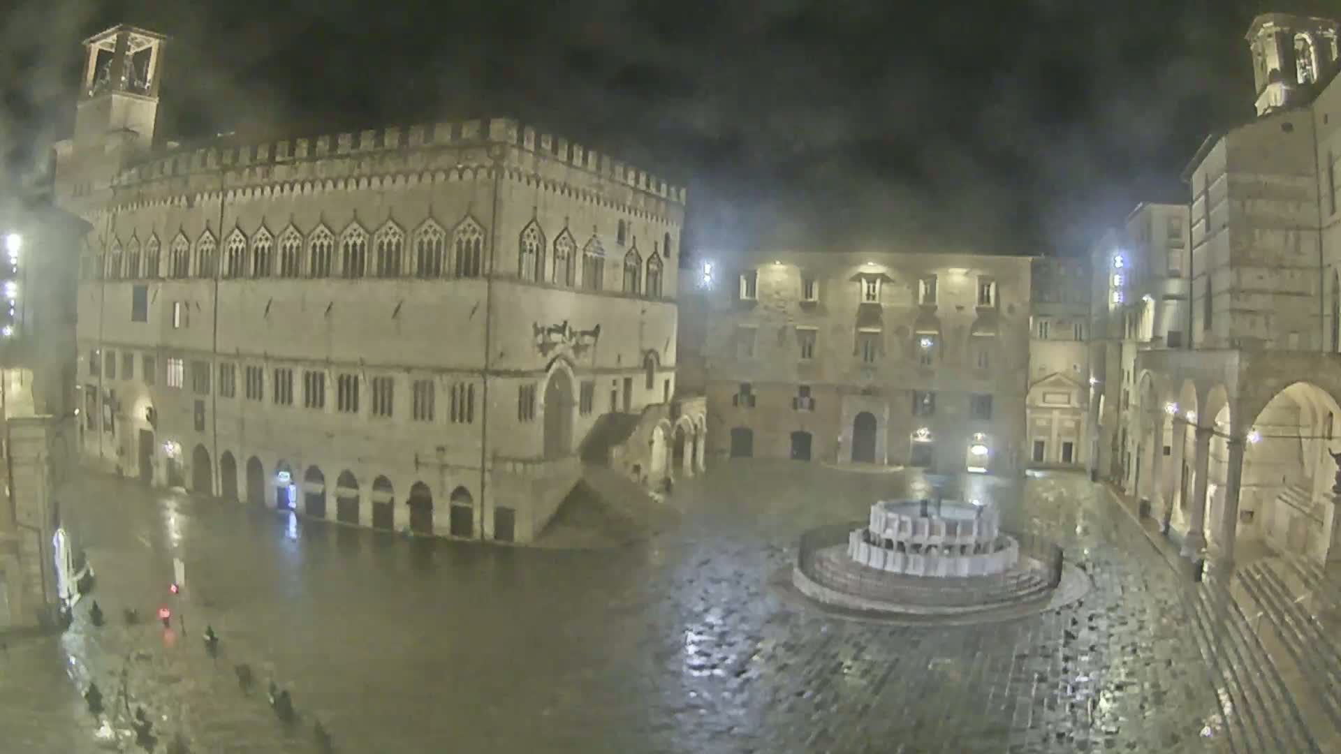 A large, stone plaza at night in the rain, centered on a fountain, is surrounded by old, multi-story buildings.
