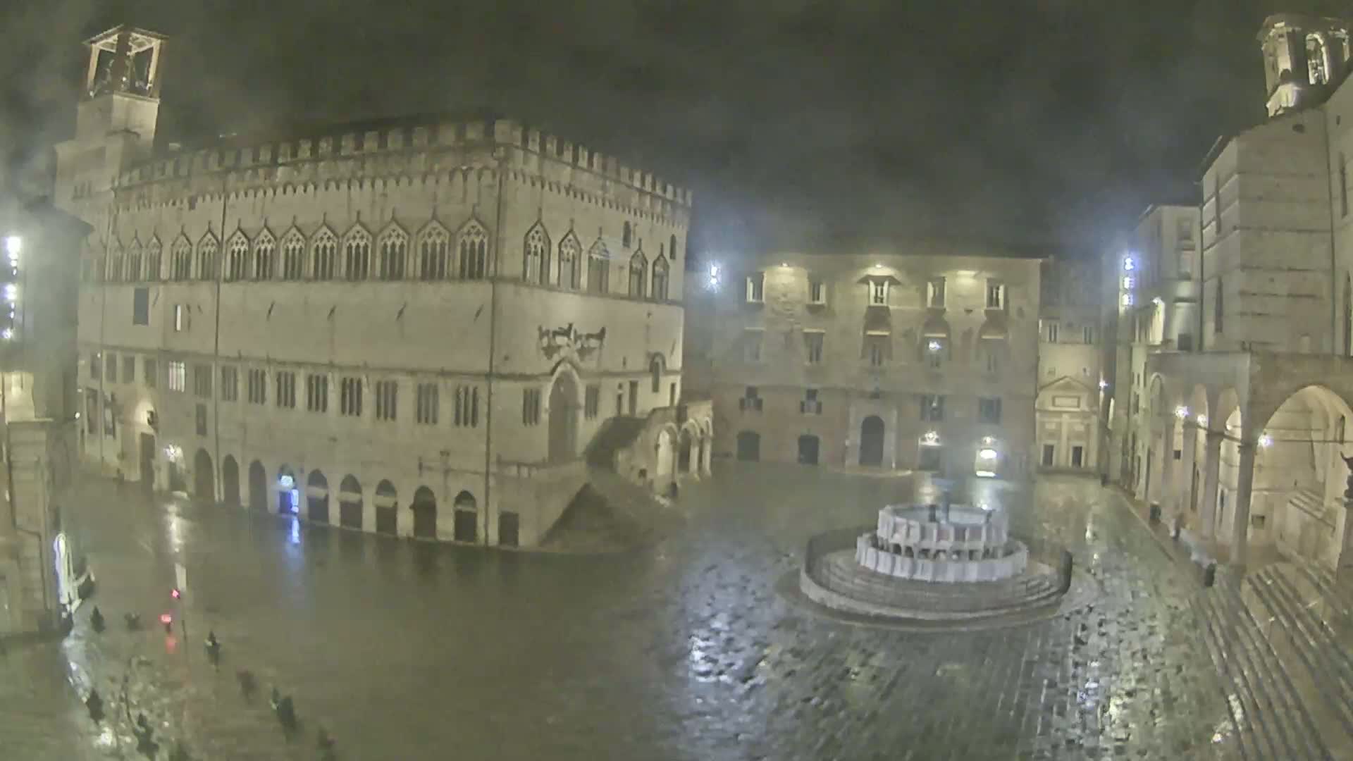 A rain-soaked town square at night, surrounded by historic buildings, with a fountain in the center.