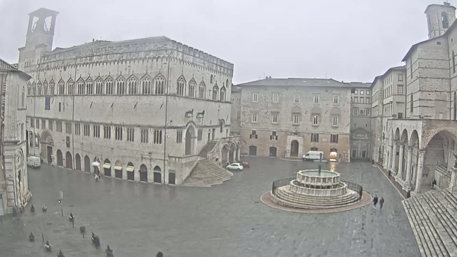 A large, stone plaza with a fountain is surrounded by ancient-looking buildings under an overcast sky.