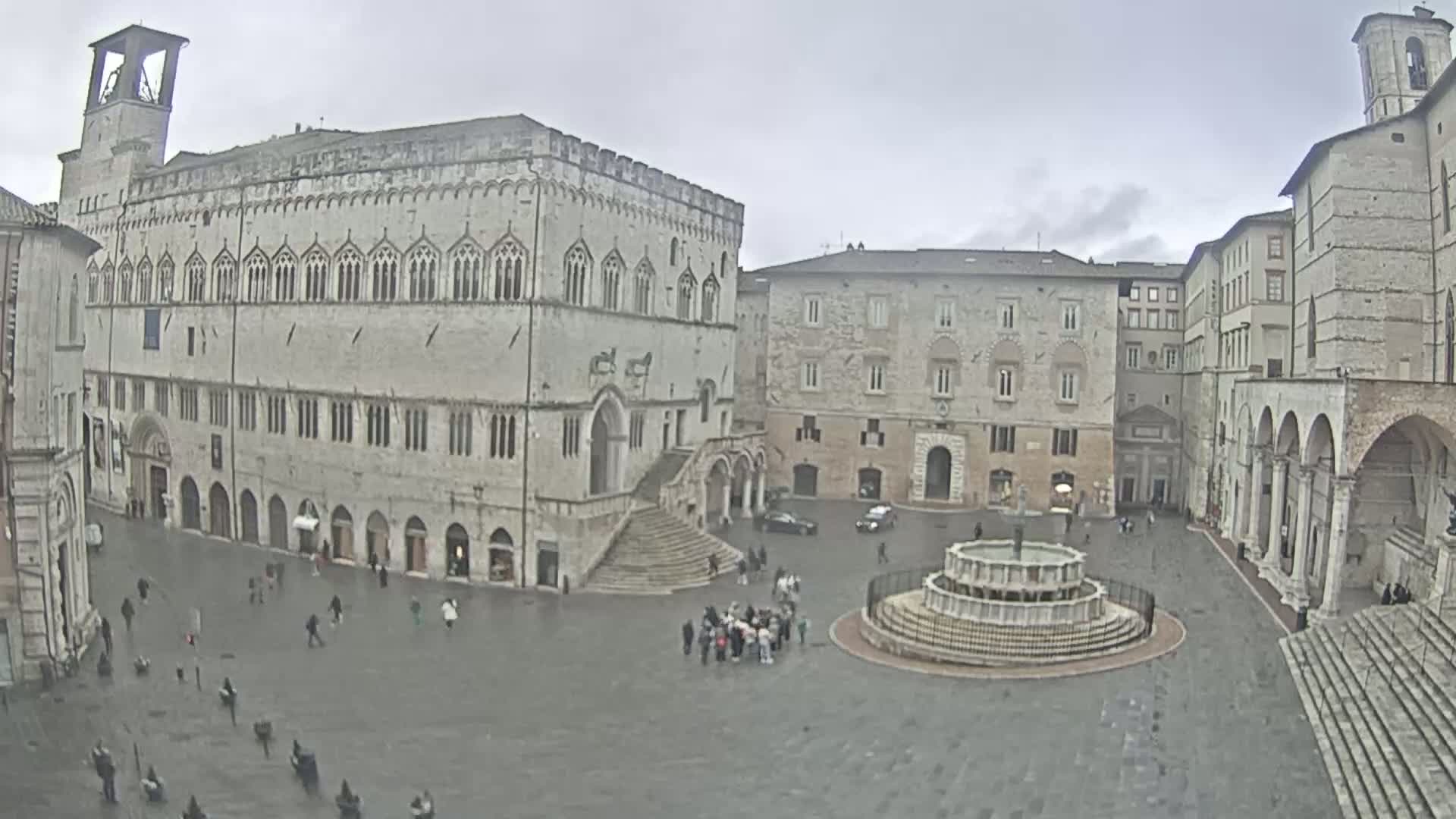 A large, stone plaza with a fountain in the center, surrounded by historic buildings under an overcast sky.