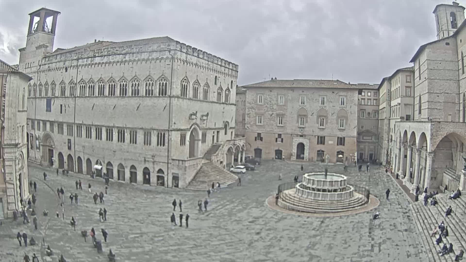 A large town square with several stone buildings, a fountain, and a scattering of people under an overcast sky.
