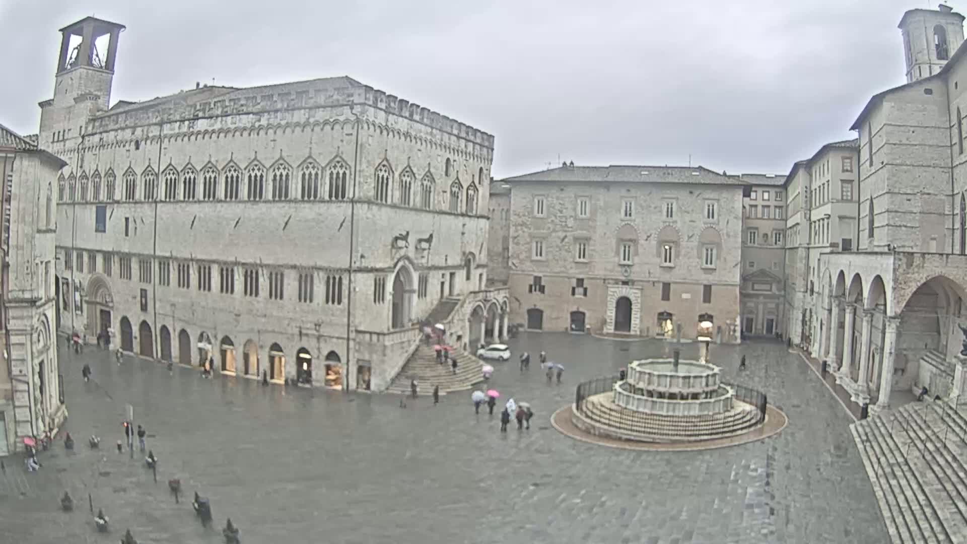 A large town square with several stone buildings, including a large palace, is shown on a rainy day, with a few people carrying umbrellas.