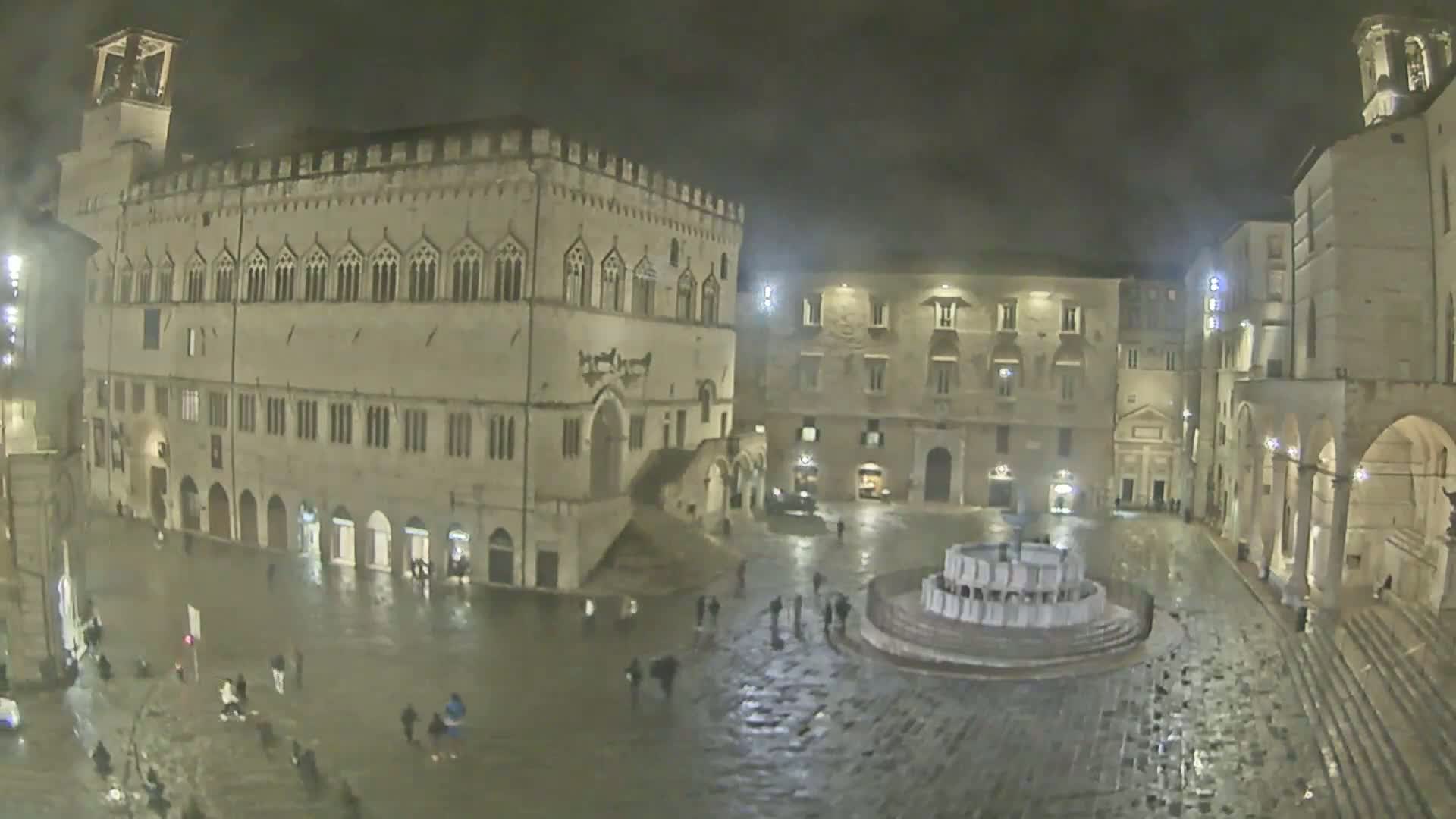 A large, stone plaza at night, illuminated by lights, is wet from rain and features a fountain in the center, surrounded by several historic buildings.