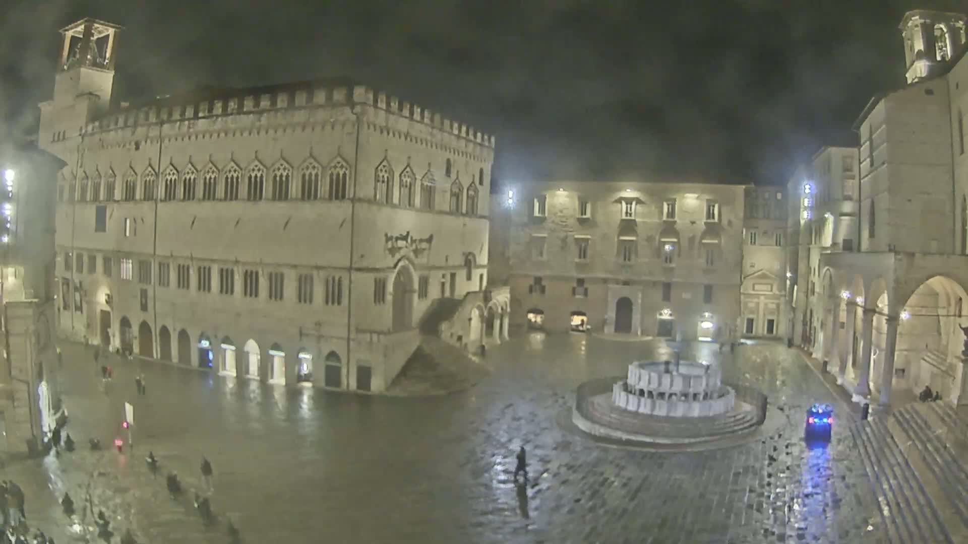 A nighttime view of a large, mostly empty town square with several historical buildings surrounding a fountain, under rainy conditions.