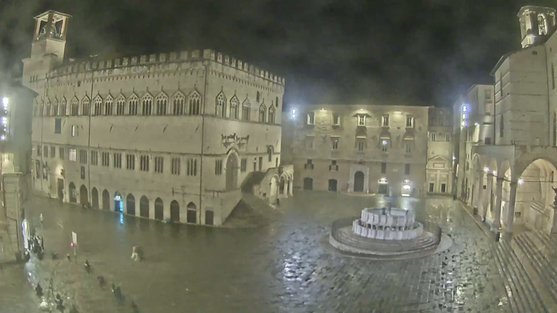 A large, empty town square at night, surrounded by old stone buildings, is partially flooded under a dark, rainy sky.