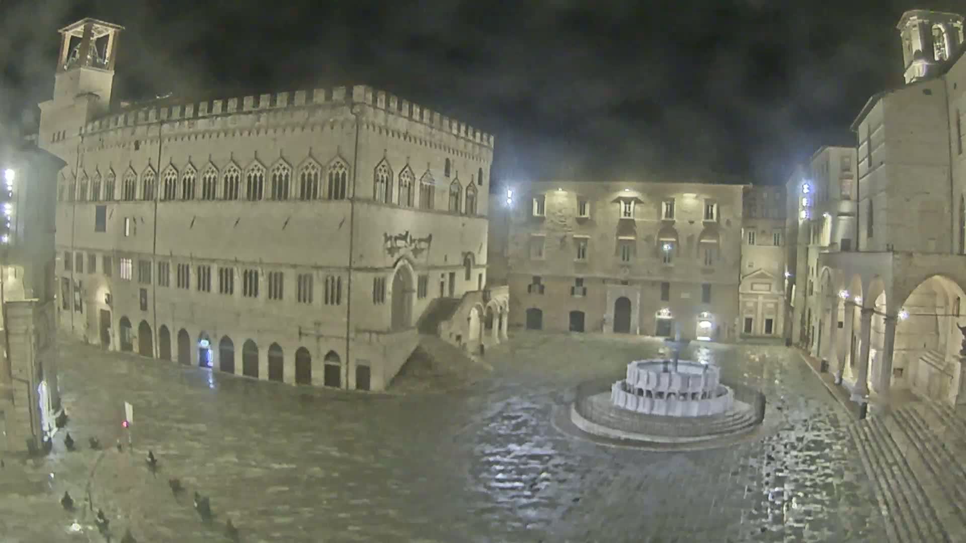 A large, stone plaza at night, illuminated by lights, is flooded with rainwater and features a central fountain surrounded by historic buildings.