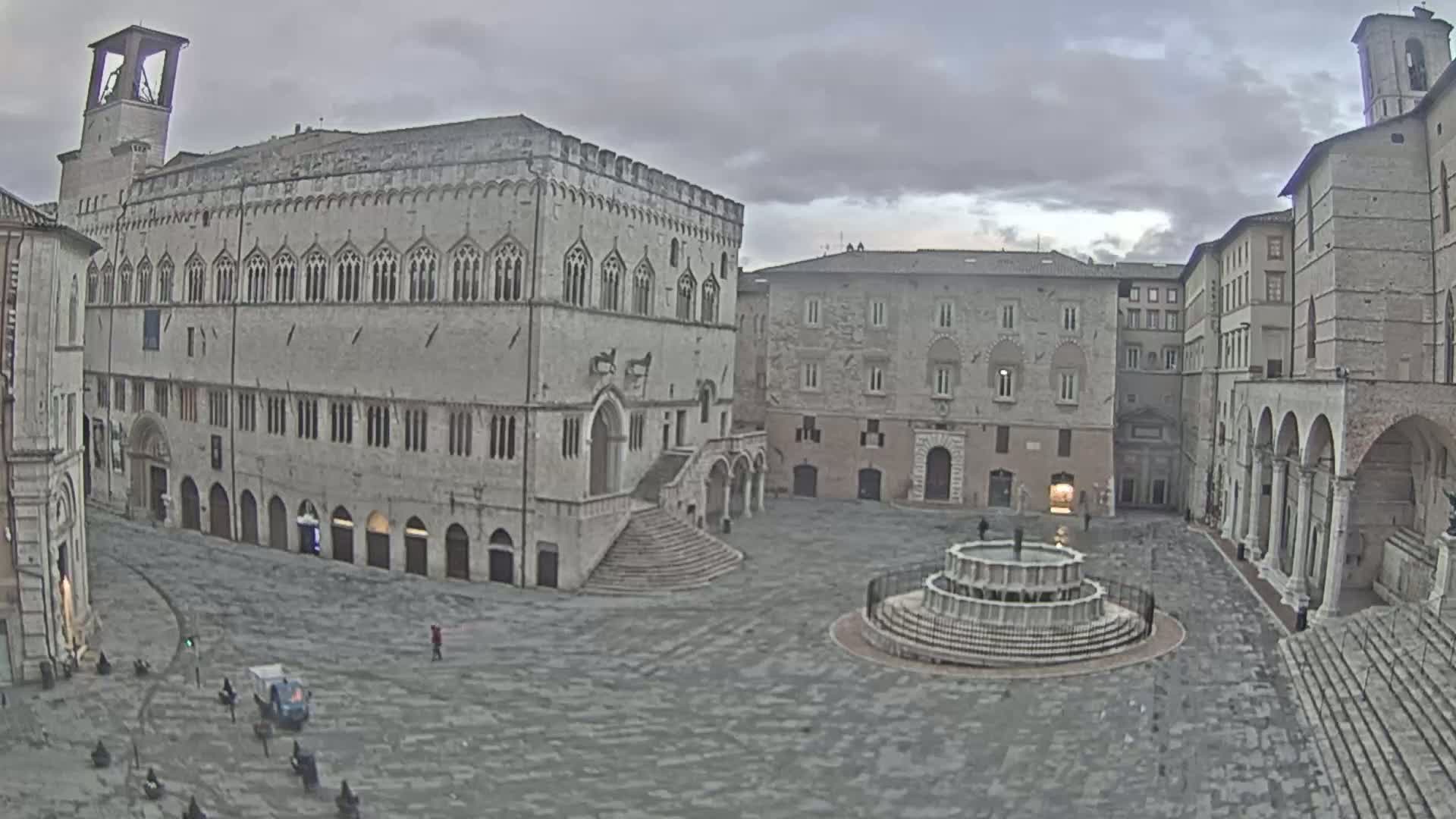 A large, mostly empty stone plaza is centered around a circular fountain, surrounded by several aged, multi-story buildings under a cloudy sky.