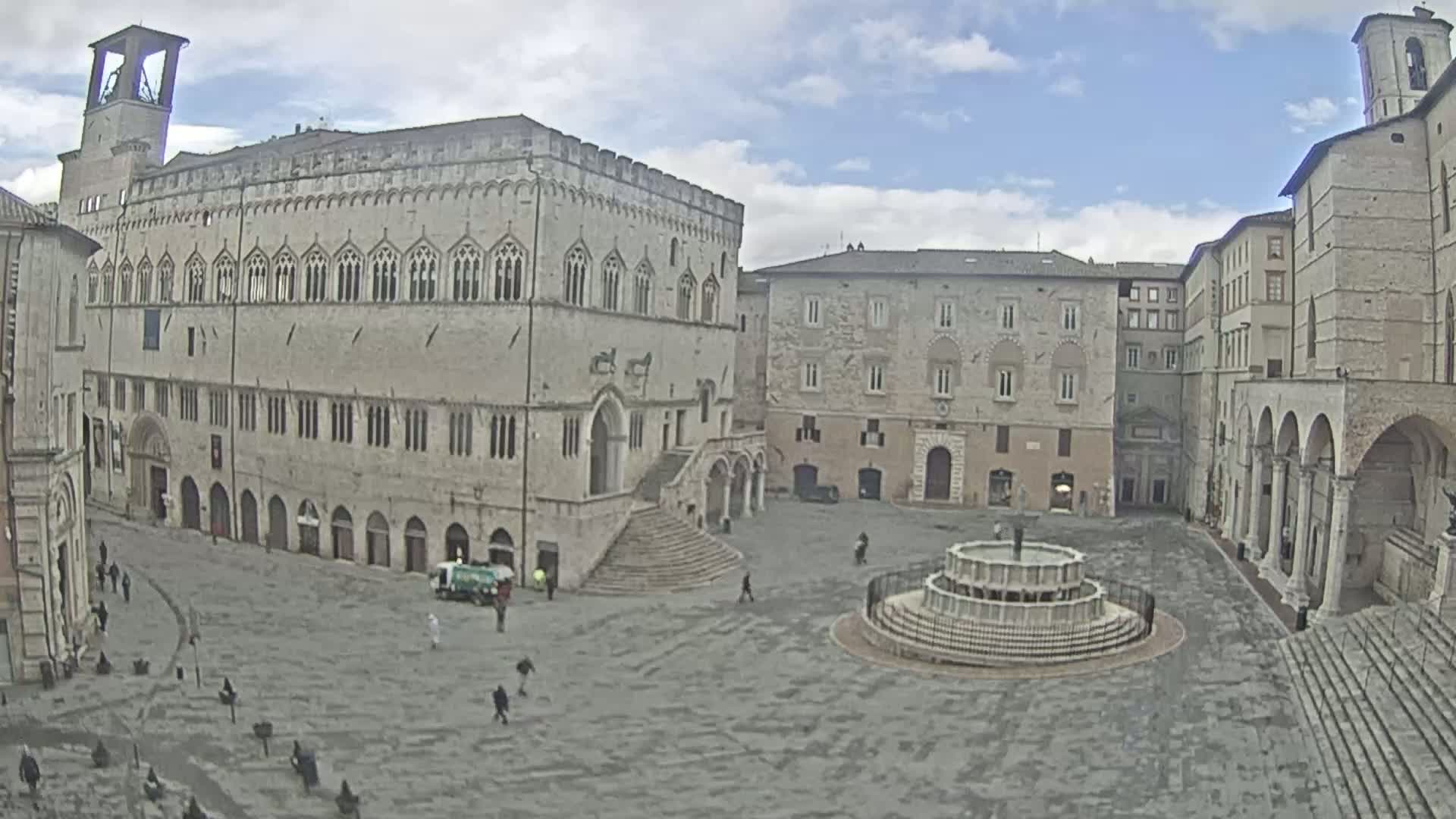 A large, mostly empty town square with a fountain in the center is surrounded by several stone buildings under a partly cloudy sky.