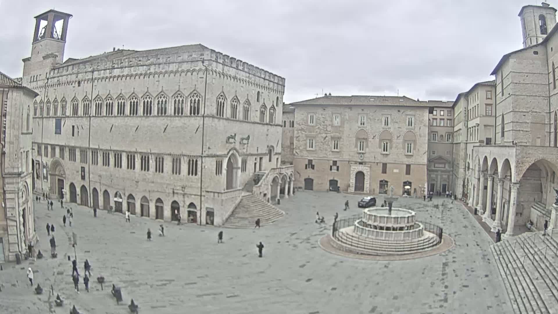 A large, mostly empty town square with several stone buildings and a fountain, under an overcast sky.