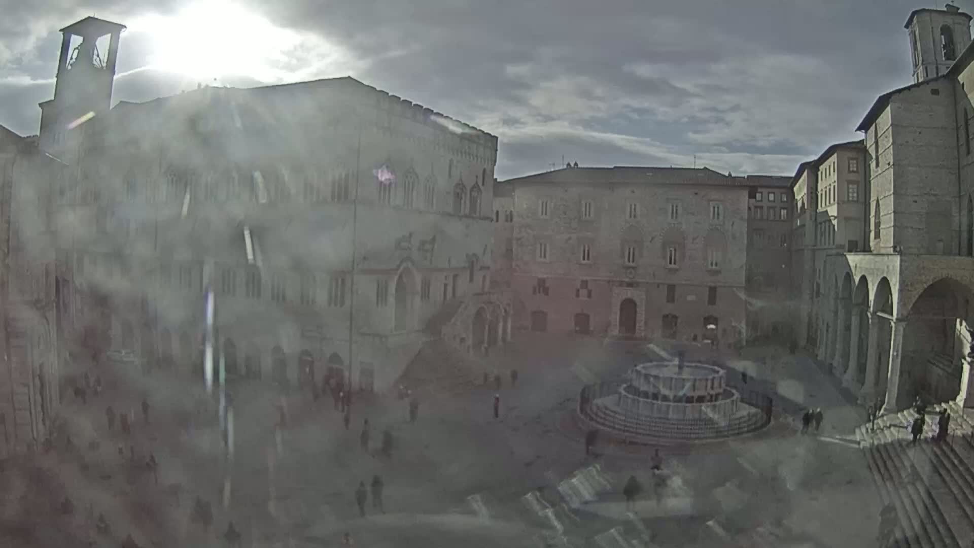 A hazy sun shines over a large, stone plaza surrounded by old, multi-story buildings, a fountain at the center.