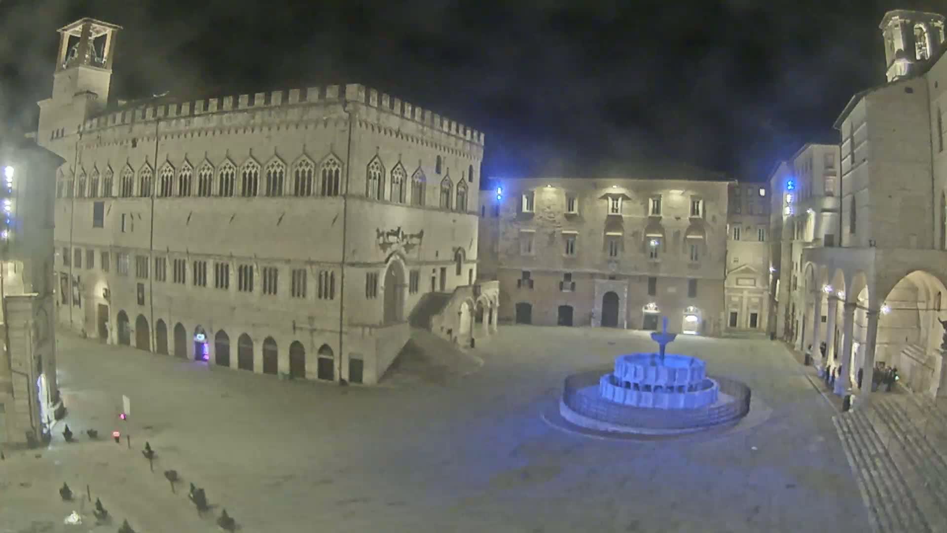 A nighttime view of a town square with a fountain, surrounded by historic stone buildings under a dark sky.
