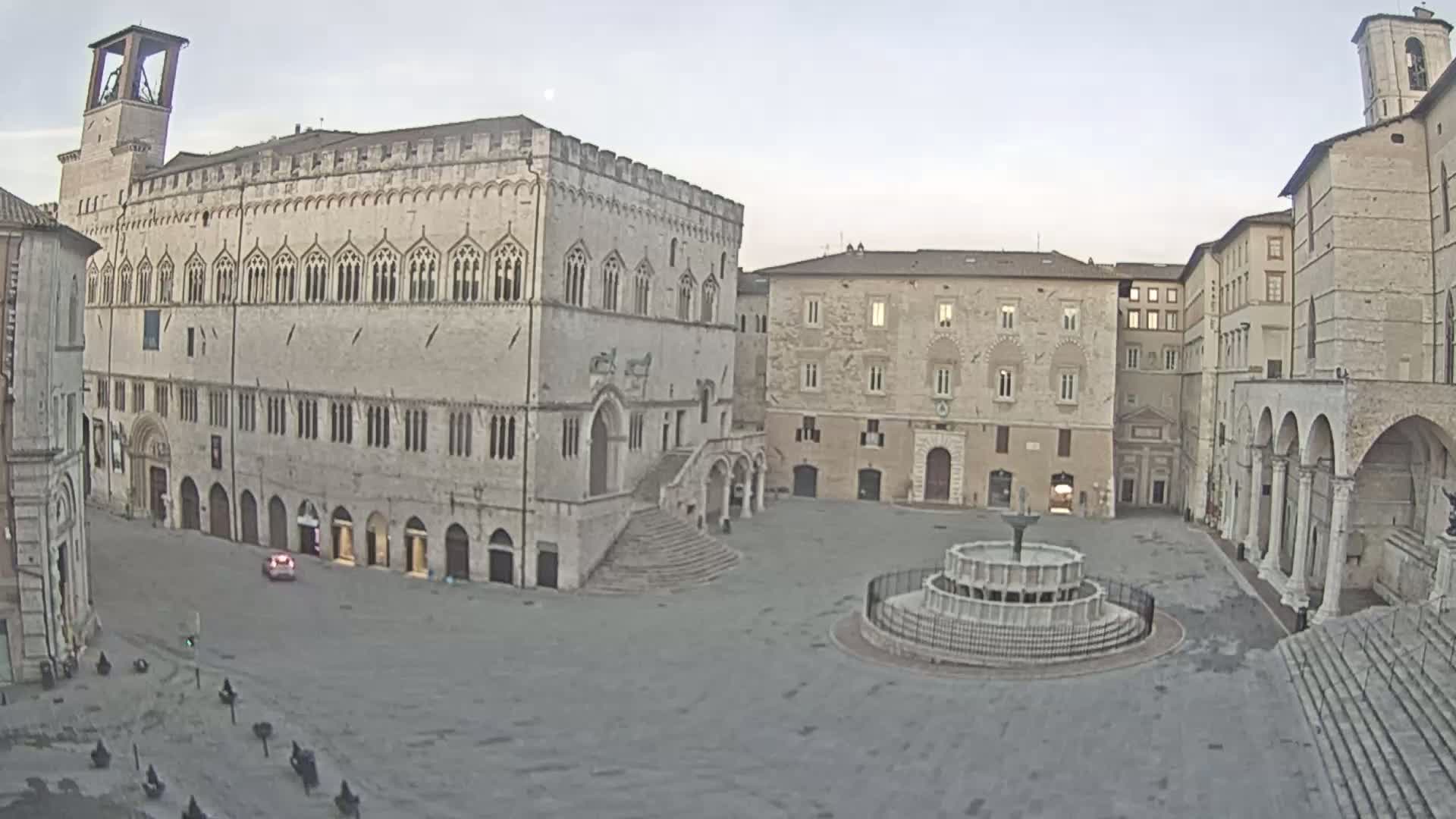 A large, mostly empty town square is surrounded by stone buildings, one of which has a prominent staircase, under an overcast sky.