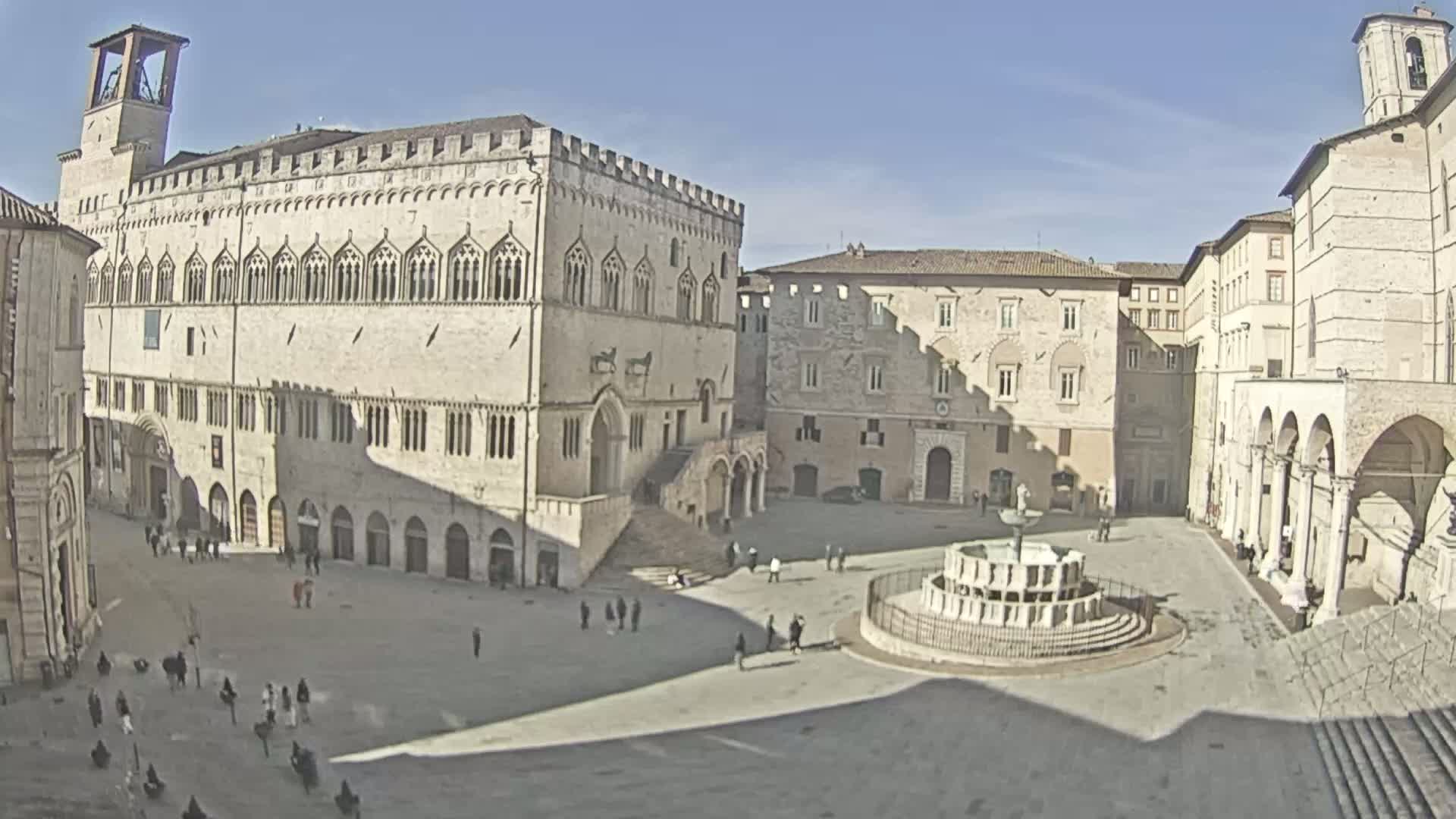 A large, sunlit town square is surrounded by historic stone buildings, featuring a central fountain and a few people sparsely scattered throughout.